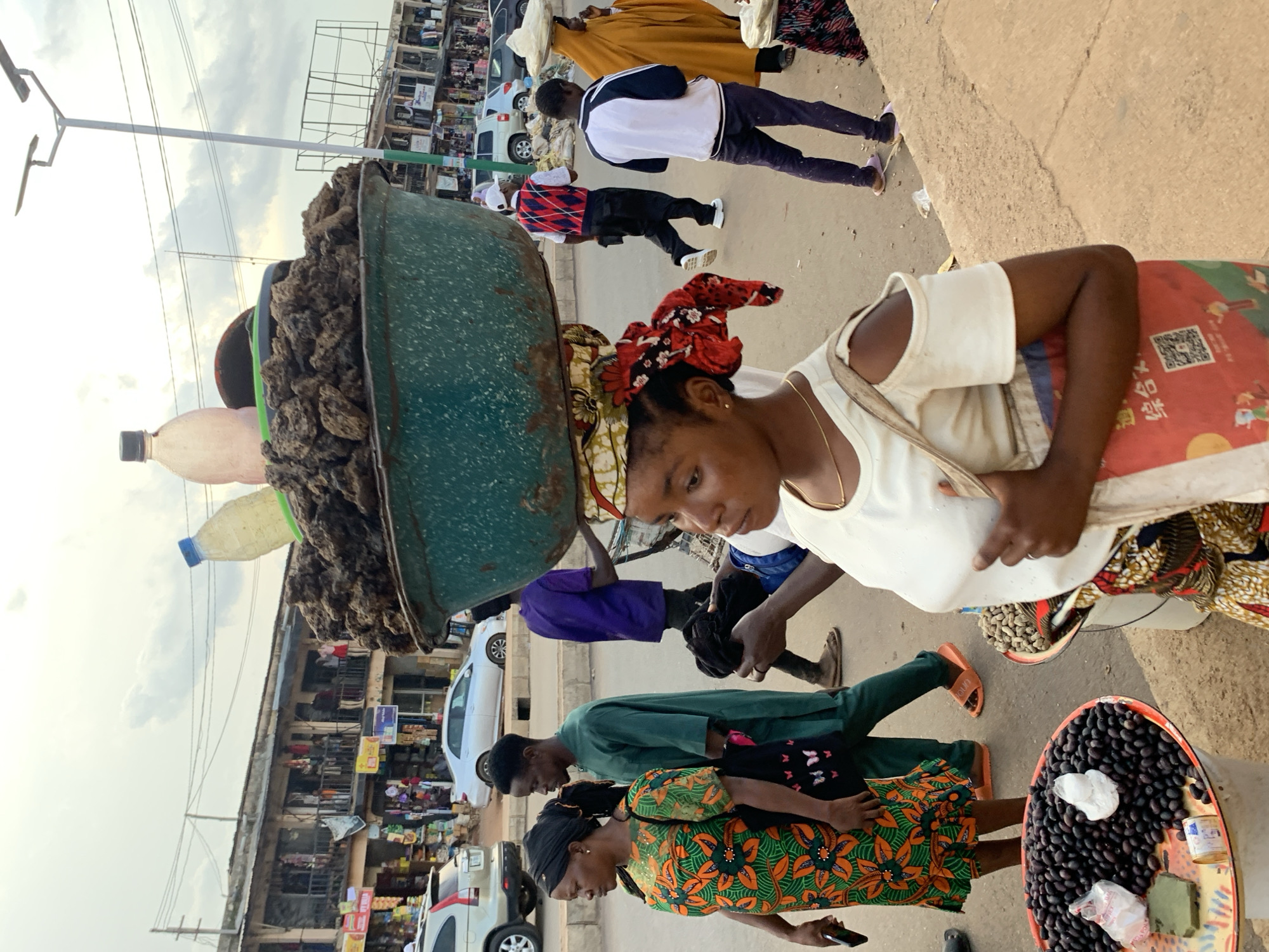 The image depicts a vibrant street market scene in Jos, Nigeria. Several people are visible, including women carrying goods on their heads and men walking through the market. One woman in the foreground is looking towards the camera, wearing a white top and a colorful patterned skirt, with a red and black headwrap. Behind her, another woman in a green outfit is walking, and a woman in an orange and green patterned dress is also present. Men are seen in the background, some wearing traditional attire.
Objects in the scene include a large green basin filled with what appears to be produce or dried goods, being carried on a woman's head. Several plastic bottles, one filled with a yellow liquid, are also visible. A cart or stall is set up, displaying a large tray of dark, round fruits, likely olives, along with some white items and small bottles. In the background, market stalls are lined up with various goods displayed, and vehicles, including cars and a motorcycle, are present on the street.
The setting is an outdoor market on a paved street, with buildings lining one side of the market. The sky is overcast, suggesting a cloudy day. The overall atmosphere appears to be one of a busy marketplace with people engaged in commerce. The presence of multiple individuals and the arrangement of goods indicate a typical day in a Nigerian market. There are no explicitly visible emotions displayed by the subjects.
There is some visible text on a bag in the foreground with Chinese characters. There are also various signs on the market stalls in the background, but the text is not clearly legible.