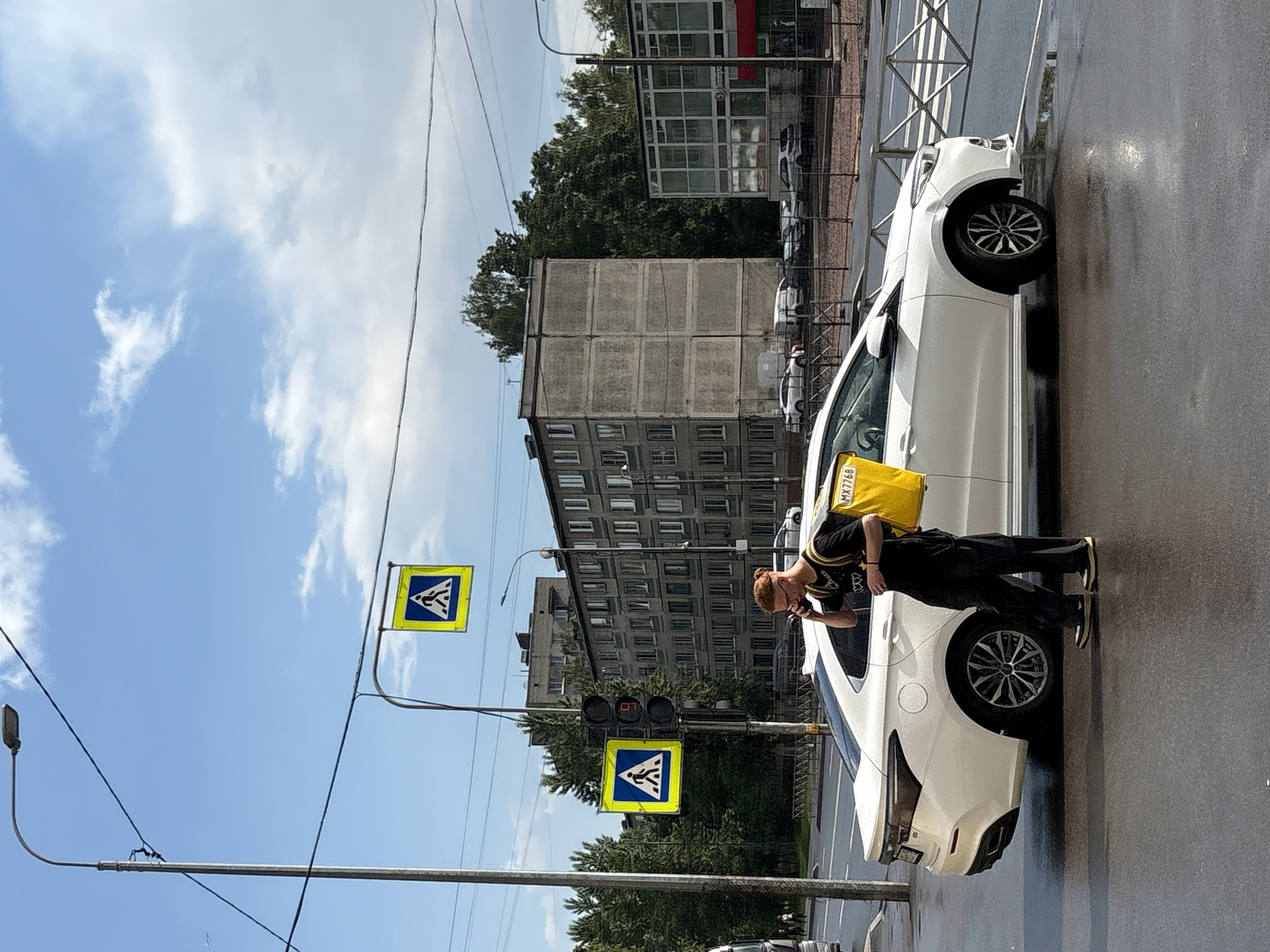 A person wearing a black t-shirt, black pants, and a yellow backpack is standing on a wet road in Saint Petersburg, Russia, talking on a mobile phone. The person is next to a white car parked on the street. In the background, there is a multi-story apartment building, a street light, and pedestrian crossing signs. The sky is blue with some clouds.