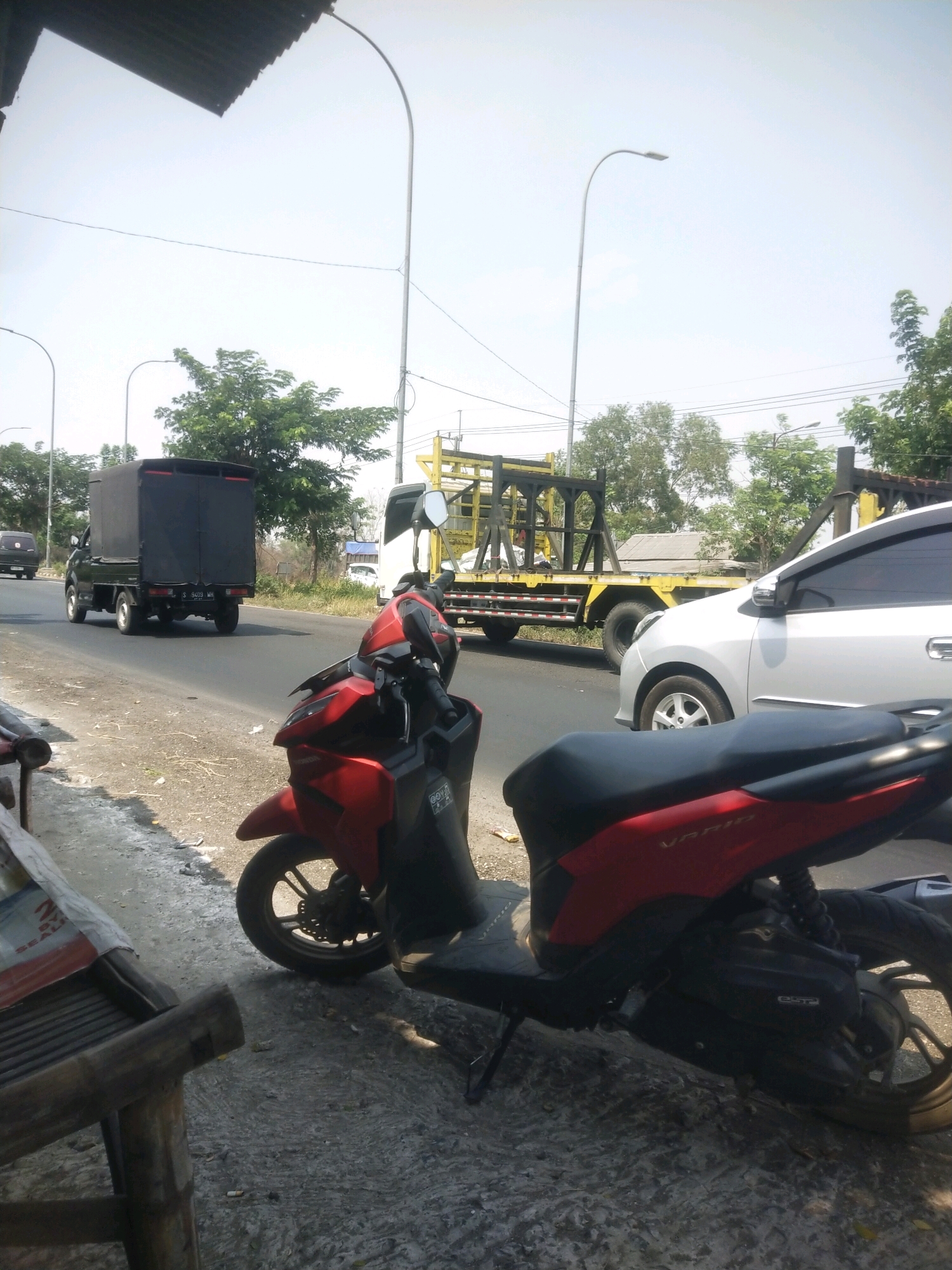 The image depicts a street scene in Petapan, Indonesia, on a sunny day. In the foreground, a red scooter is parked, facing right. The scooter has a black seat and dark gray engine casing. To the left of the scooter, a portion of a wooden bench is visible, covered with a white tarp. The ground is a mixture of asphalt and concrete.
In the midground, a paved road runs horizontally. Several vehicles are present on the road. A black pickup truck with a covered bed is driving away from the viewer on the left side of the road. Further down the road, a yellow flatbed truck with a construction frame is driving towards the viewer. Next to the flatbed truck, a silver car is also driving towards the viewer.
The background features a clear blue sky and lush green trees. Streetlights are visible on the left and right sides of the road, with one positioned near the center. The overall environment suggests a typical urban or semi-urban setting in Indonesia. No people are clearly visible in the image.