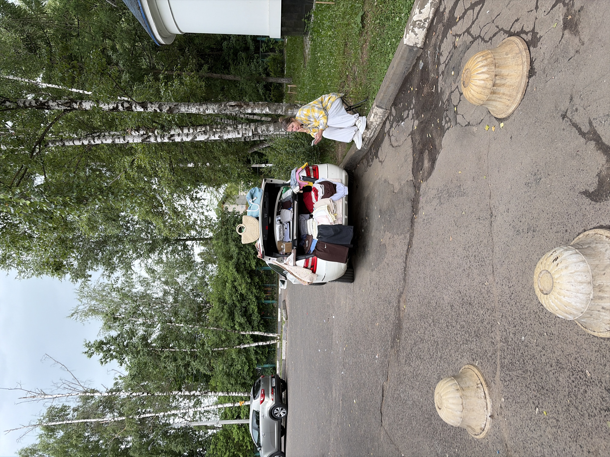 A woman sits on a folding stool on the side of a paved area, looking at her phone. She is wearing a yellow plaid jacket and white pants. Beside her, a white car has its trunk open and is packed with clothing, bags, and boxes, suggesting a move or a large shopping trip. The car is parked on asphalt, and a silver SUV is visible in the background. The environment includes a grassy area with trees and a white building with a curved roof. Two decorative, bell-shaped concrete planters are in the foreground on the asphalt. The scene appears to be in a residential area of Пикино, Russia, given the presence of birch trees and the architectural style of the building. The sky is overcast.