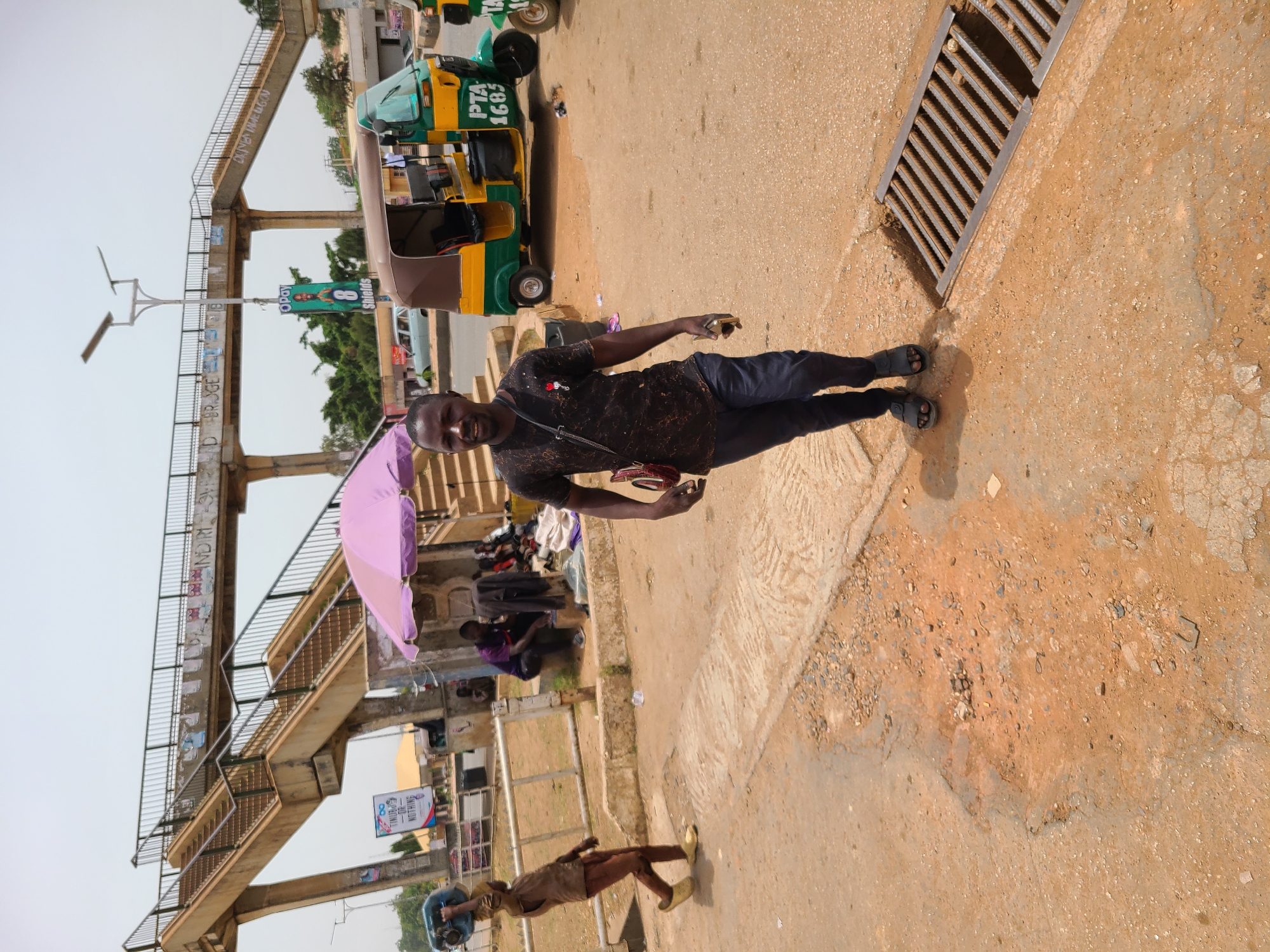 This daylight image captures an outdoor urban scene in Jos, Nigeria, characterized by pedestrians, local transport, and market activity. A man with dark skin, wearing a patterned dark shirt, dark trousers, and dark sandals, stands in the foreground, looking at the camera with a slight smile. He holds a small red object and a phone.
To his right, a yellow and green three-wheeled auto-rickshaw (keke napep) is parked, bearing the text "PTA 1683" on its side. A large metal storm drain grate is visible on the ground nearby. To the man's left, a vibrant purple umbrella is open, under which a person is seated, suggesting a street vendor or market stall. Further back on the left, another person in light clothing carries a large blue basin on their head.
A concrete pedestrian bridge with metal railings dominates the background, spanning across the scene. Faint graffiti, including fragmented text like "BR.DGE" and "DU...N...N", is visible on its structure. Under the bridge, various goods are spread out, and an "Opay" banner hangs, displaying the "Opay" logo and text that appears to include "8 Sh...ds". The ground consists of reddish-brown dirt mixed with cracked concrete, indicating a busy, well-trodden area. The clear sky and distinct shadows suggest a sunny day, likely late morning or afternoon.