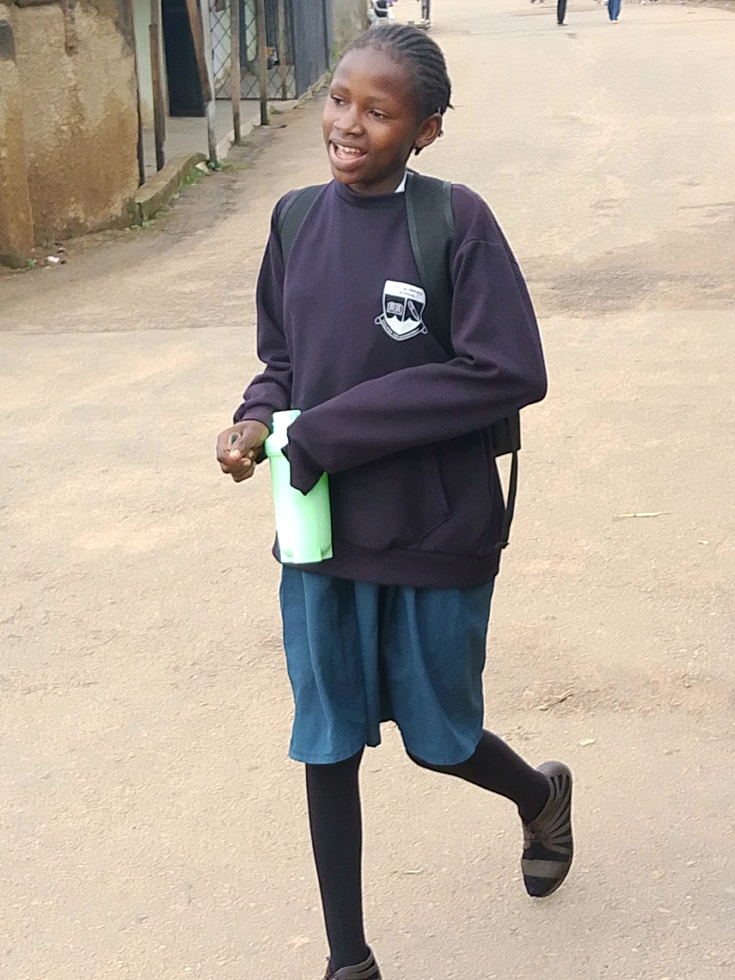 A young Black girl with braided hair, wearing a dark purple school sweater with a crest, a blue skirt, black tights, and striped shoes, walks on a dirt road in Jos, Nigeria. She carries a light green water bottle in her right hand and a black backpack on her shoulders. Buildings line the left side of the road.