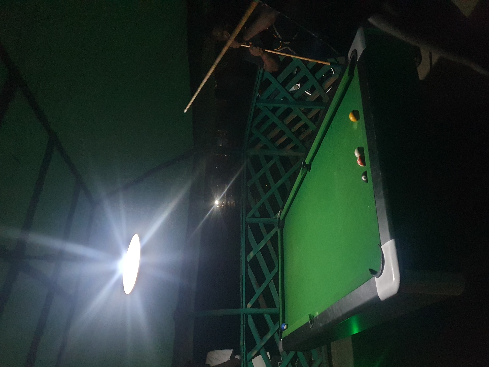 This media file shows a person playing snooker in an indoor setting at night. The scene is dimly lit by a bright overhead light that casts strong rays, creating a dramatic effect. A green snooker table is central to the image, with several balls visible on its surface, including a yellow ball, a red and white striped ball, and a blue and white striped ball. A person is partially visible, holding two snooker cues, seemingly in the act of playing. The background features a dark, possibly rustic or industrial structure with green metal latticework. The overall ambiance is that of a casual, perhaps even slightly gritty, recreational space. Given the mention of Lokoja, Nigeria, and the nighttime setting, it suggests an indoor recreational activity common in the region. There is no visible text in the image.