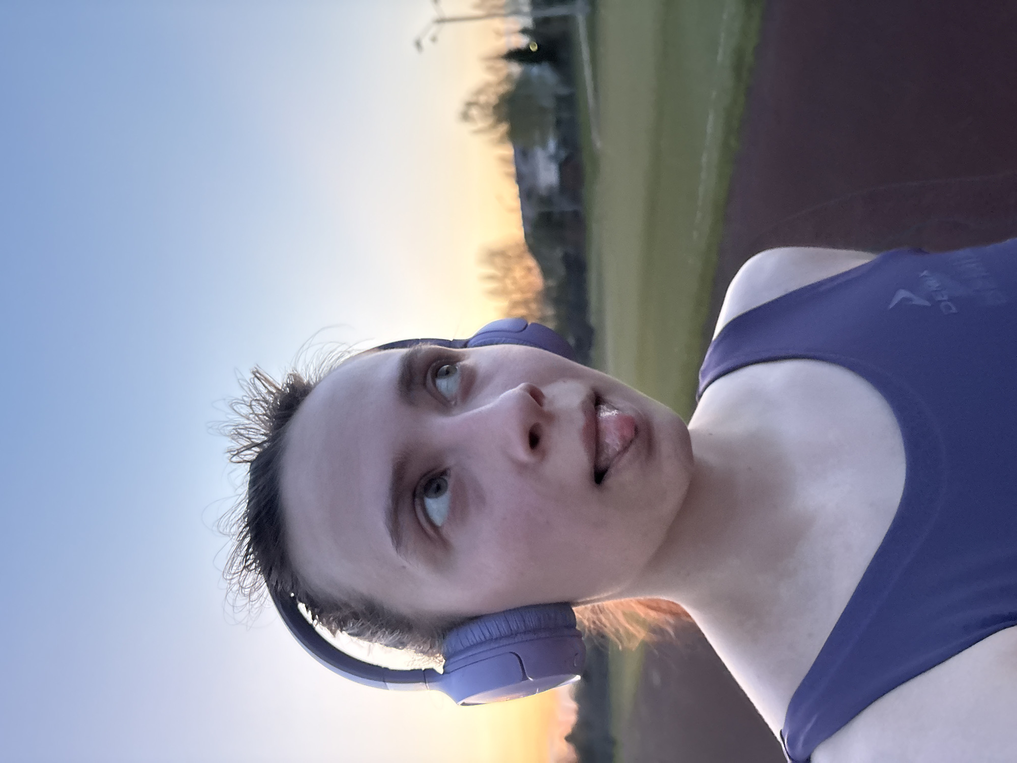 A young woman wearing a blue tank top and purple headphones looks upwards with her tongue slightly protruding. She is outdoors in a park or recreational area. The sky behind her has colors suggesting sunset or sunrise, with a mixture of blue and orange hues. There's a running track and some trees visible in the background. The location is inferred to be Прикамский, Russia, based on context.