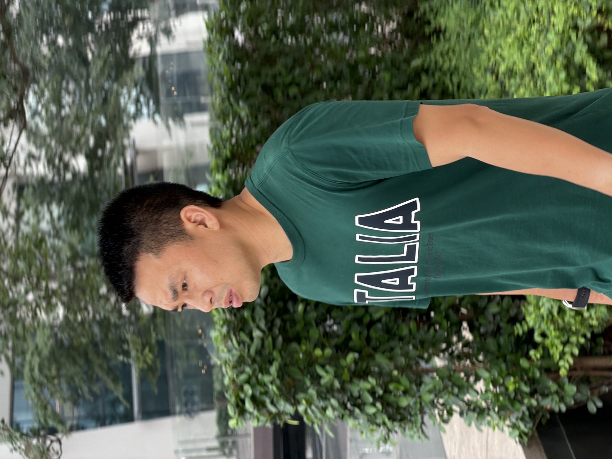 A medium shot shows a man in profile, walking outdoors in front of a blurred background of greenery and modern buildings. The man has short, dark hair and is looking to his left with his mouth slightly open. He is wearing a dark green t-shirt with white lettering that appears to read "ITALIA". The environment suggests an urban setting, possibly a park or a landscaped area outside of a commercial building. The lighting indicates it is daytime.
