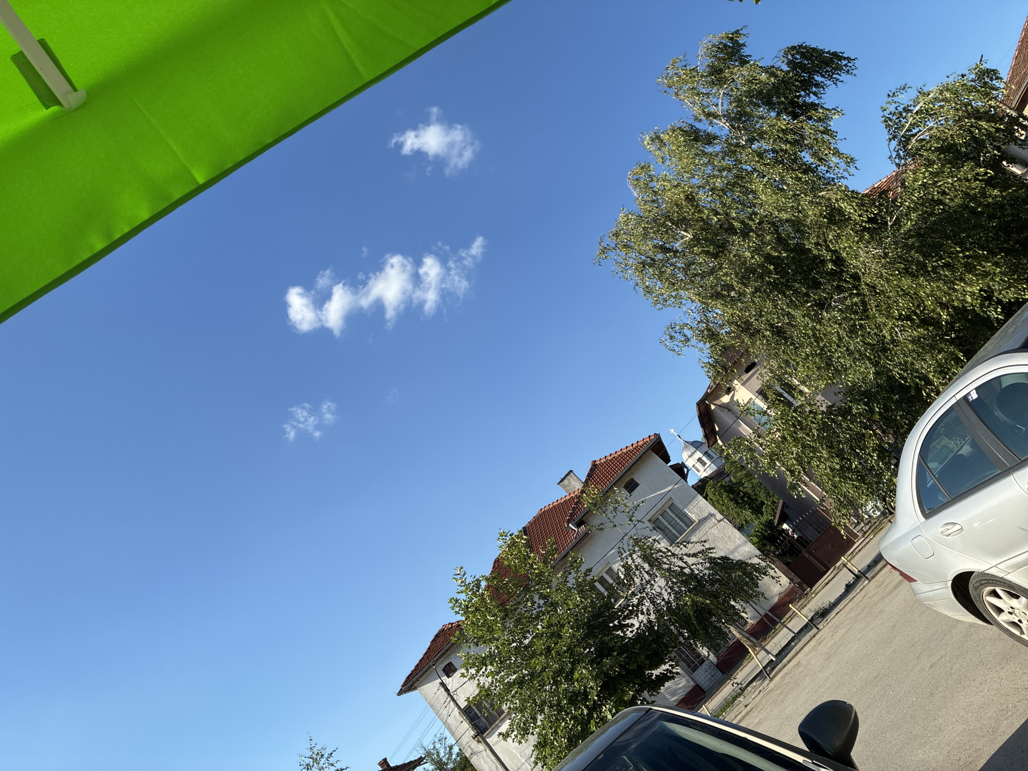 In Aglen, Bulgaria, on a clear day with a bright blue sky and sparse white clouds, a street scene unfolds. The perspective is tilted, as if the camera is angled upwards. A bright green canopy, possibly from an umbrella or awning, dominates the upper right portion of the frame. Below it, a street is visible, lined with buildings and trees. Two cars are parked on the street: a dark-colored vehicle on the left and a silver sedan in the foreground. Trees with dense green foliage line the street, their branches reaching upwards. The buildings appear to be residential, with red tiled roofs. One building features a white facade and several windows. A small church or tower with a cross is partially visible between the trees. The overall impression is a quiet, sunlit day in a residential area.