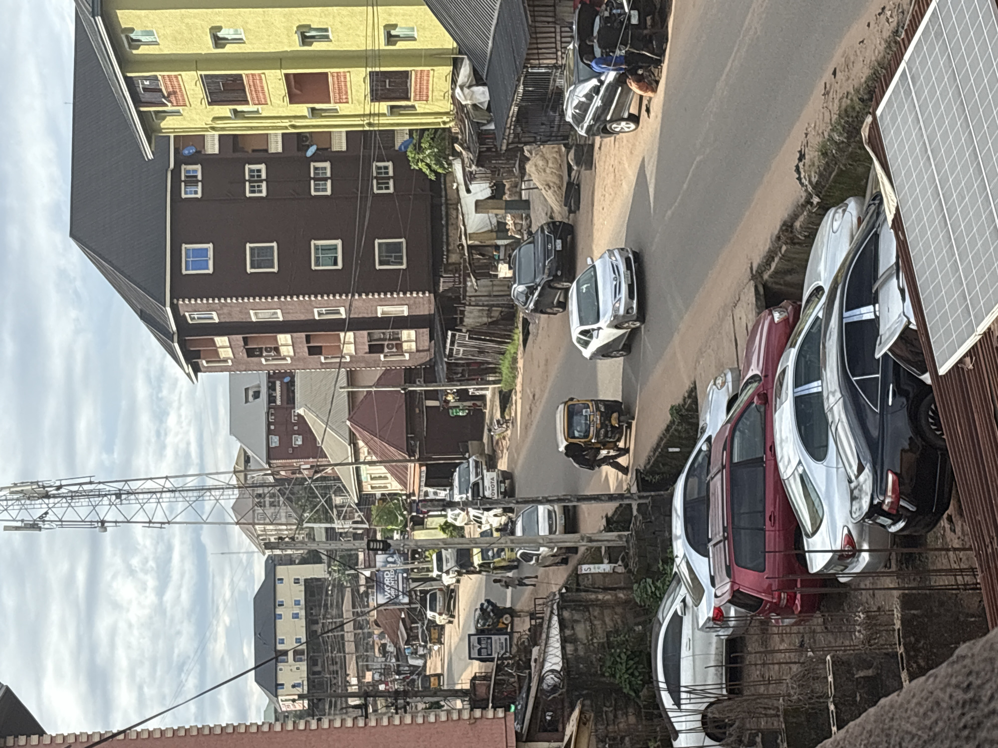 The image depicts a street scene in Nkpor, Nigeria, likely a commercial or residential area. The foreground is dominated by the roof of a structure with corrugated iron and a solar panel installation. Beyond this, several cars are parked or moving on a paved road. Vehicles include a black sedan, a silver sedan, and a red SUV, with other cars partially visible further down the street.
The midground shows a bustling street with a variety of vehicles, including cars, a white Toyota pickup truck, and a yellow auto-rickshaw (Keke Napep). People are visible interacting with some of the vehicles. A person is standing next to the auto-rickshaw, and another individual is walking with a load on their head. Buildings line the street on both sides. These include multi-story residential buildings with balconies and windows, some painted in brown and yellow. There are also smaller structures with tin roofs and open-front shops. A prominent cell phone tower stands tall in the background, suggesting proximity to telecommunications infrastructure. The sky is overcast with clouds. The overall impression is of a busy, everyday urban environment in Nigeria.