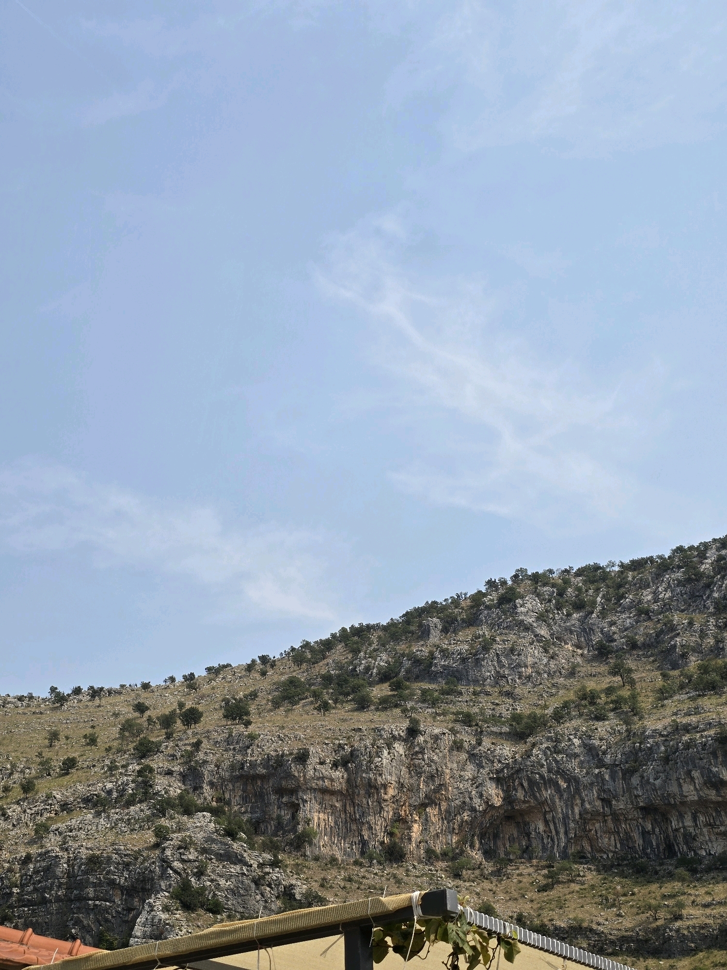 The image shows a rocky hillside with sparse vegetation under a clear blue sky with wispy clouds. The hillside is covered in grey rocks and green shrubs, with some larger trees scattered across its surface. At the bottom of the image, there is a section of a building with an orange-tiled roof and a tan-colored awning or overhang. This appears to be a natural landscape with some human structures. The location is described as being in the city of Podgorica, Montenegro, which suggests this is a mountainous or hilly area on the outskirts of the city. There are no people or animals visible in the image, and no actions or interactions are taking place.