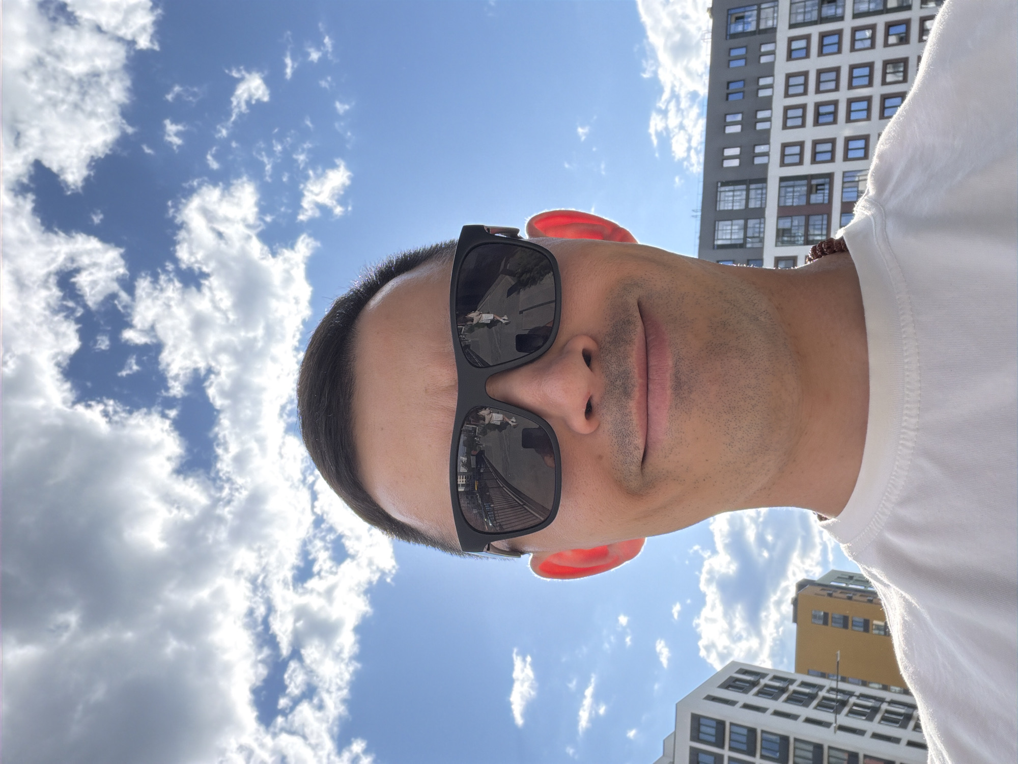 A selfie-style portrait captures a male subject from the chest up, centered in the lower portion of the frame, against an urban backdrop in Izhevsk, Russia. The man has short, dark hair, wears dark rectangular sunglasses, and a white t-shirt, with a dark, possibly beaded, necklace partially visible on his right side. He exhibits a faint smile. Bright sunlight illuminates his face, particularly noticeable through his left ear which appears reddish. The reflections in his sunglasses reveal a street scene: the left lens shows a paved surface alongside a barrier or fence with buildings in the distance, while the right lens prominently reflects a female pedestrian, dressed in a light top, shorts, and white sneakers, walking away from the viewer on a similar paved path. The background behind the man features a bright blue sky with scattered white cumulus clouds, and several modern multi-story buildings, including a white facade building on the left, a dark grey building on the right, and a yellow building further back between them. The overall scene indicates a clear, sunny day.