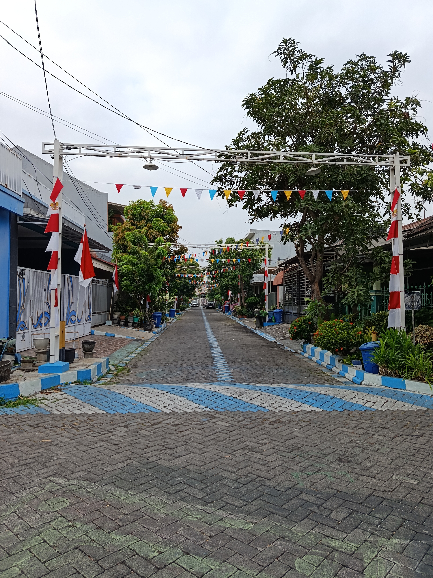 The image depicts a street in Keboansikep, Indonesia, adorned with festive decorations. Red and white Indonesian flags are prominently displayed, along with colorful triangular flags strung across the street. The street itself is paved with bricks, featuring a blue and white patterned design. Houses line both sides of the street, with lush green trees and plants contributing to the environment. The sky is overcast, suggesting a cloudy day. There are no people visible in the image.