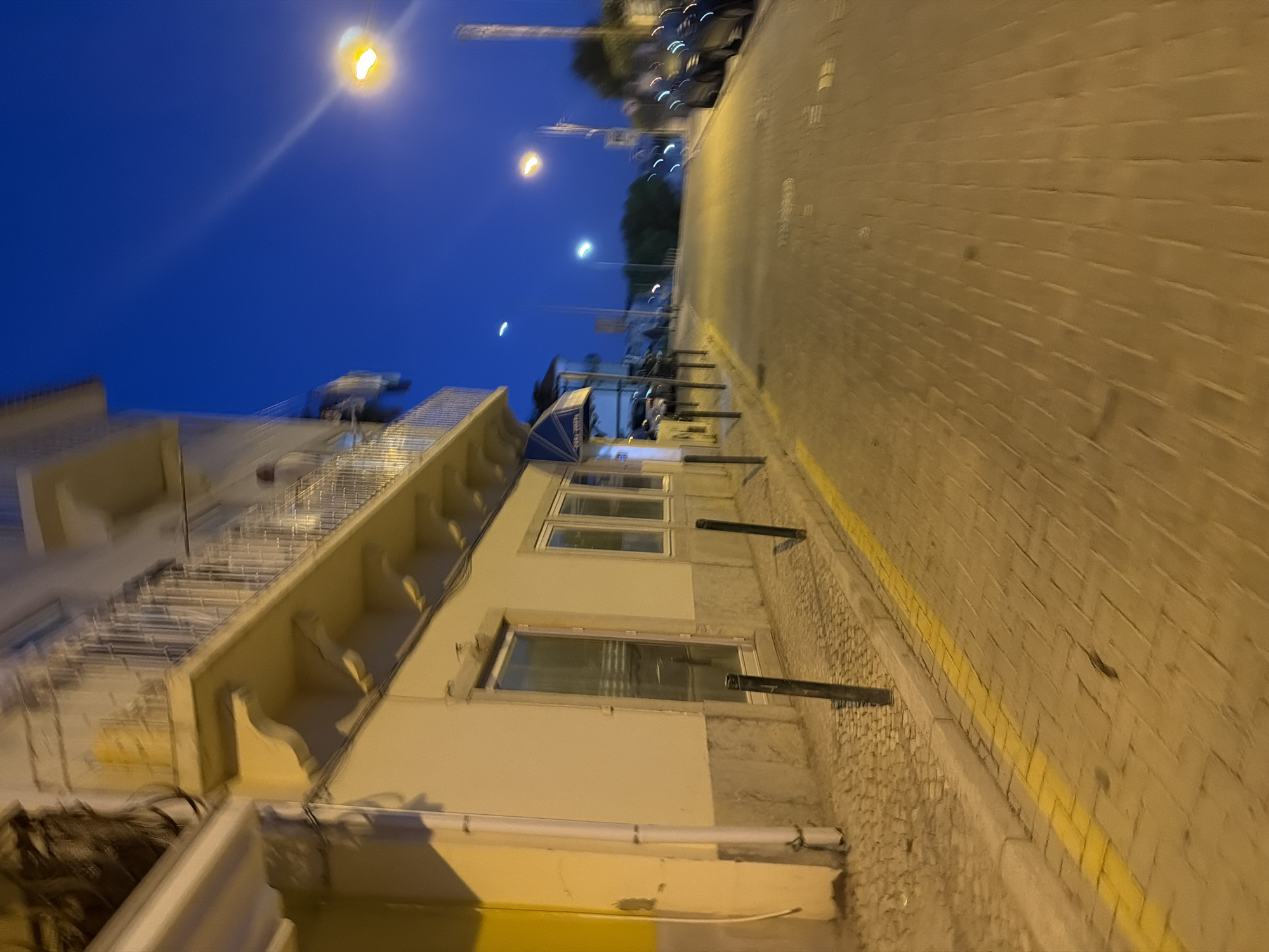 The image depicts a street scene in Cascais, Portugal, at dusk or night. The perspective is from street level, looking down a cobblestone street lined with buildings on the left and parked cars on the right. The sky is a deep blue, with visible streetlights casting a warm glow.
On the left side of the image, a light-colored building features a balcony with a metal railing. The lower part of the building has windows and a doorway. A white drainpipe runs vertically along the building's exterior. The sidewalk in front of the building is paved with cobblestones and has a bright yellow line along its edge. Black bollards are spaced along the sidewalk.
The street itself is paved with cobblestones and stretches into the distance. On the right side of the street, several cars are parked, their lights reflecting on the wet pavement. Streetlights are visible at intervals along the street, with one prominent streetlight in the upper right corner of the image. The overall impression is a quiet, residential or commercial street illuminated by artificial light against a darkening sky.