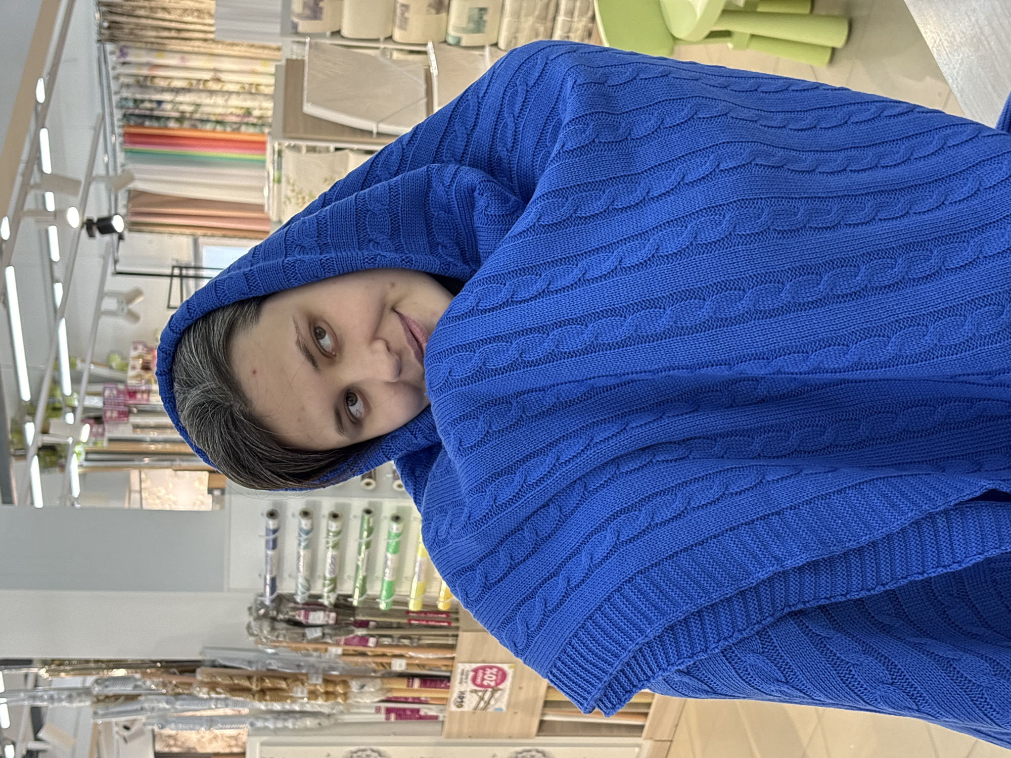 A woman with gray hair and pale skin looks at the camera while wrapped in a blue, knitted shawl or blanket. The setting is a retail store, likely specializing in textiles or fabrics. Shelves filled with rolls of fabrics and other materials are visible in the background. A "20%" discount sign is also visible. The location is in the city of Chaikovsky, Russia.
