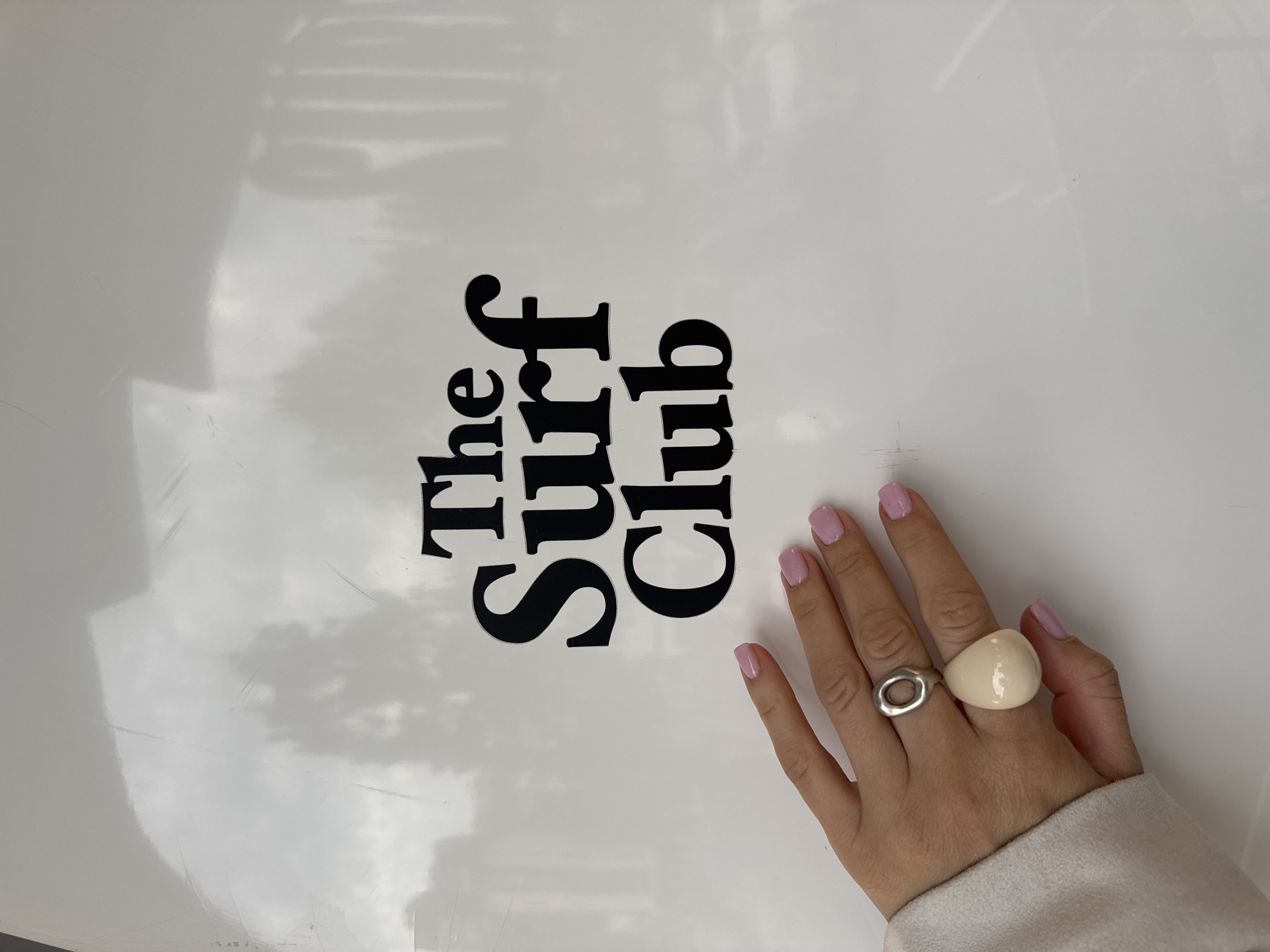 A close-up shot shows a female hand with two rings resting on a white surface with black lettering. The lettering reads "The Surf Club" in a vertically stacked arrangement. The hand has light pink nail polish on the fingernails and is adorned with two rings: one with an oval cutout and another large, round, cream-colored ring. The background is a bright, diffused white with a subtle reflection of what appears to be a cloudy sky. The bottom edge of the frame shows a small portion of what seems to be a light-colored fabric. There is no indication of the location being Sochi, Russia.