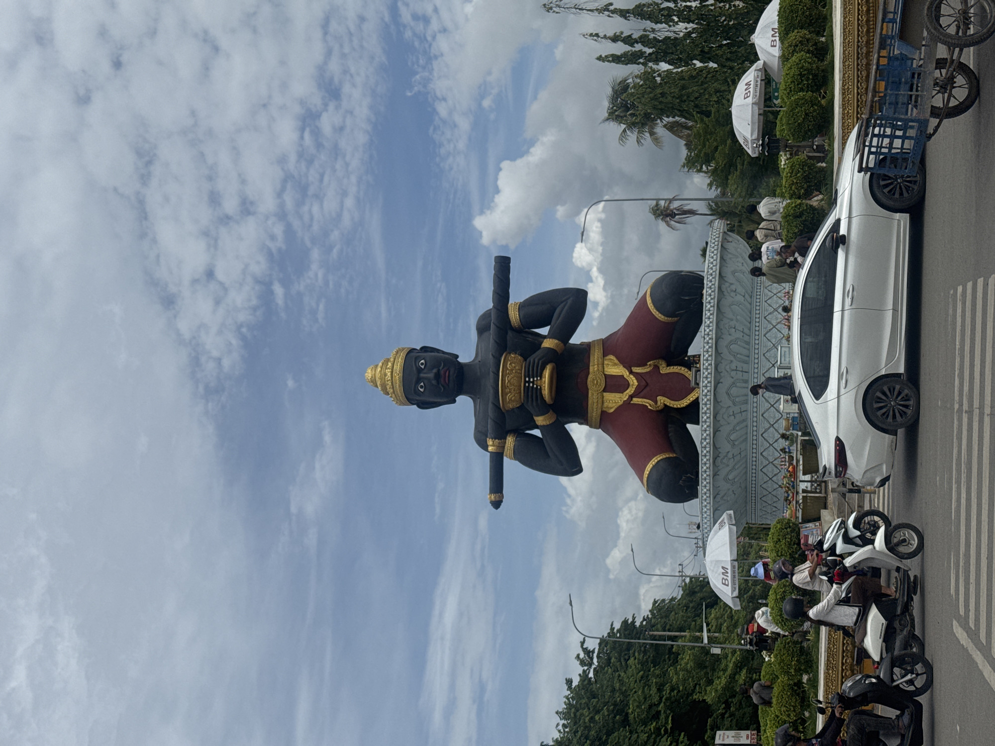 The image shows a large, dark statue of a seated figure wearing a gold headdress and elaborate gold and red clothing. The figure holds a golden bowl in its hands and has a long, dark cylindrical object resting on its shoulders and held by its arms. The statue is positioned on a large, ornate blue and white base. In the foreground, there is a street with a white sedan driving on it. Several people are visible around the statue, some standing near it and others on motorcycles. There are also white umbrellas with a "BM" logo scattered around the base of the statue. The environment appears to be a public square or roundabout in Battambang, Cambodia, with trees and streetlights visible in the background. The sky is cloudy.