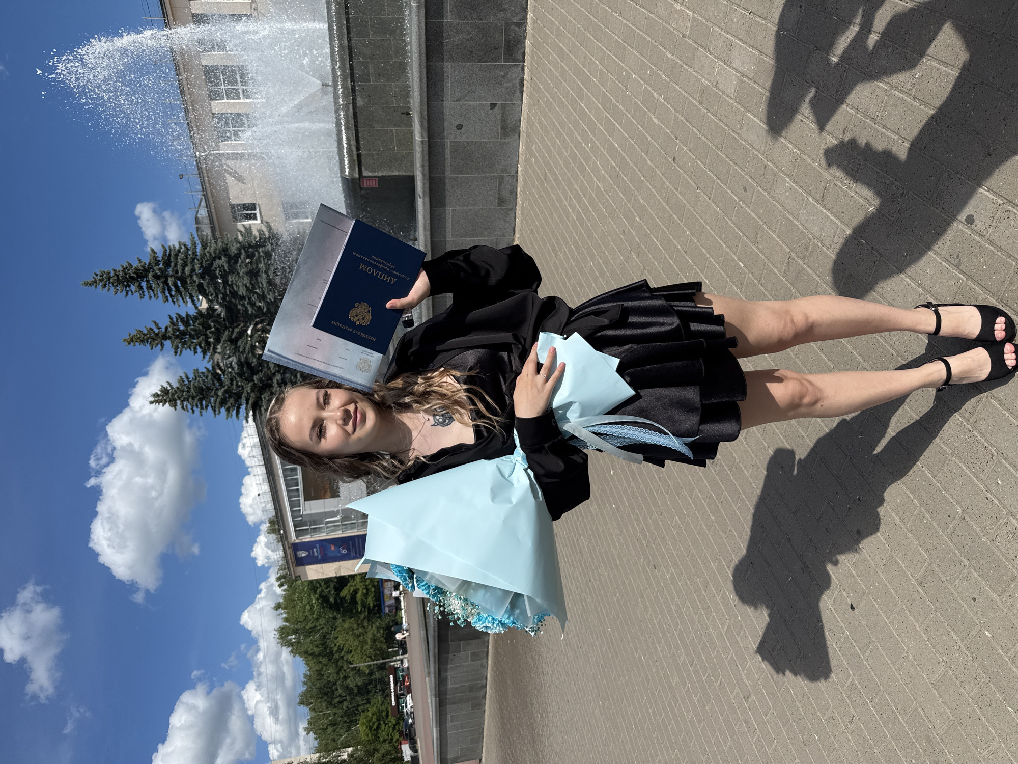 In Chaikovsky, Russia, a young woman stands outdoors, holding a diploma and a bouquet of light blue flowers wrapped in paper. She is wearing a black dress and black high-heeled shoes. Behind her, a large fountain sprays water in the air, and a building with windows and trees is visible. The sky is blue with white clouds. The ground is paved with grey bricks. A shadow of the woman is cast on the ground.