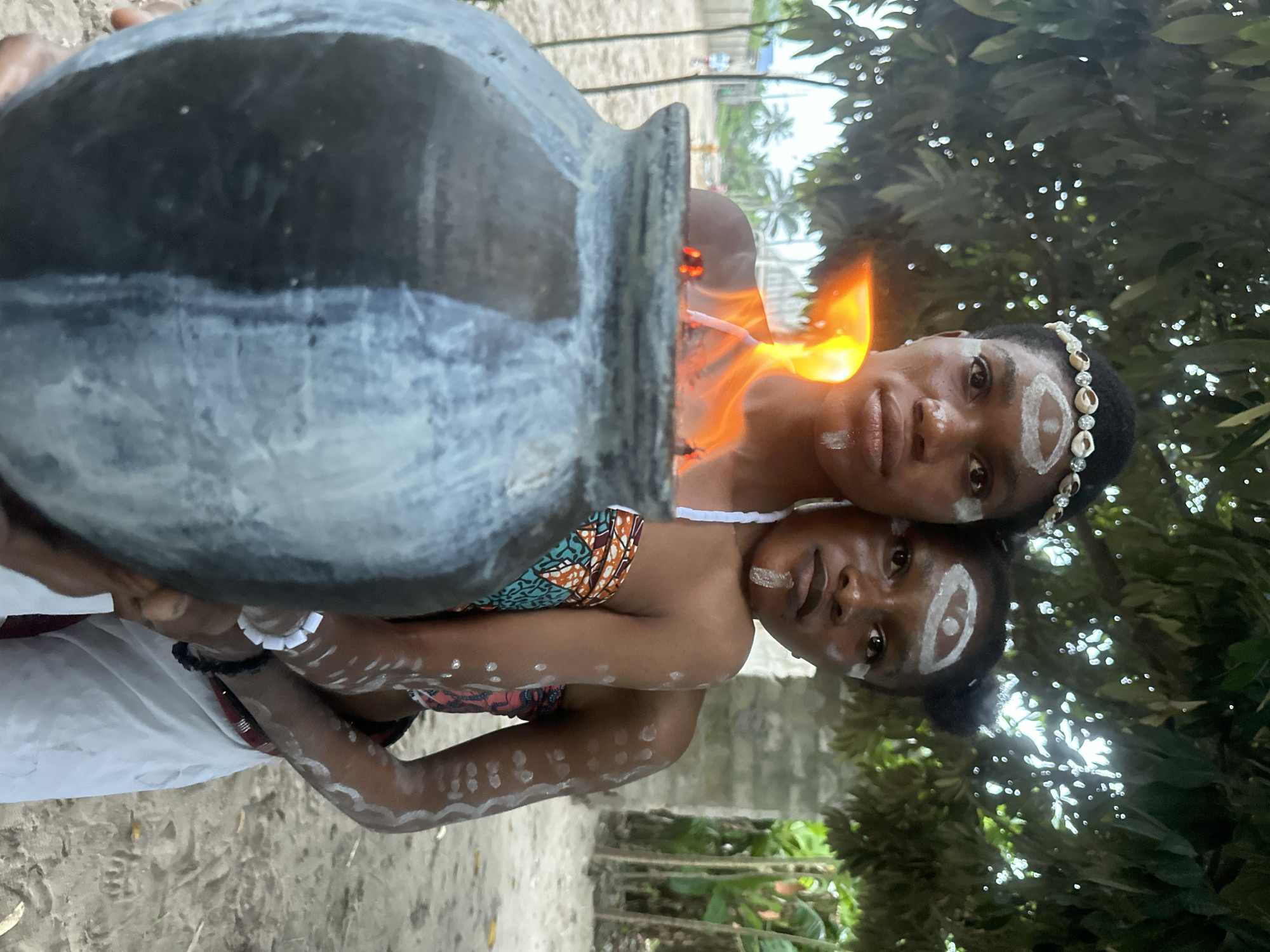 Two young women, adorned with white face and body paint and shell headbands, stand in front of a large, weathered clay pot. Flames erupt from the opening of the pot, casting an orange glow on their faces and upper bodies. The woman in front is closer to the camera, her arms wrapping around the pot, while the woman behind her has her arms around the first woman's shoulders. They appear to be standing on a sandy surface, with lush green foliage and trees in the background. A structure, possibly a building, is visible to the right. The scene suggests a cultural or ritualistic context, potentially within the historic town of Badagry, Nigeria, known for its rich cultural heritage and traditional practices.