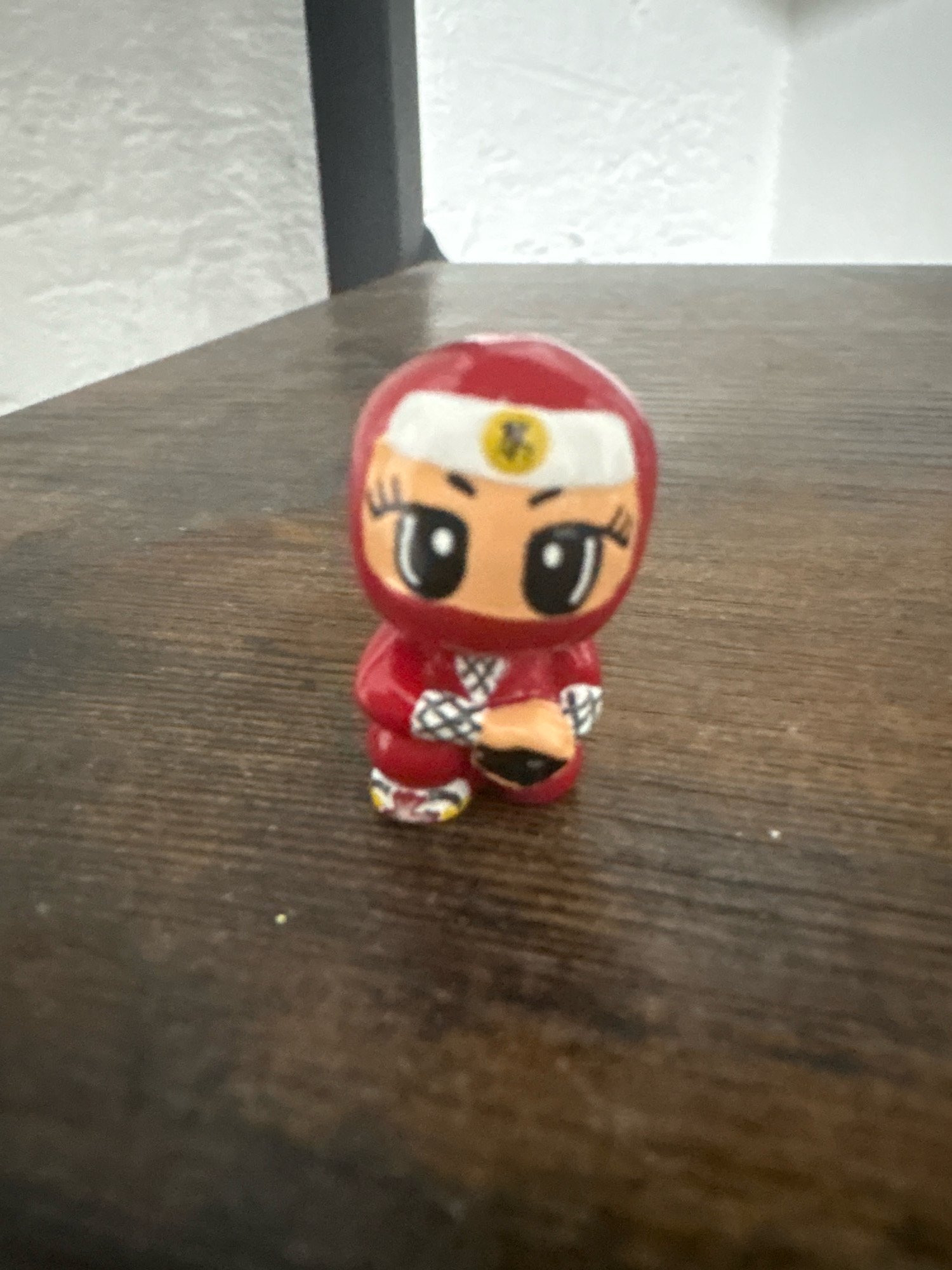 A small, red, cartoonish ninja figurine is positioned on a dark, wooden surface. The figurine has a white headband with a yellow emblem, large black eyes, and a determined expression. It wears a red outfit with white accents and holds a black object in its hands. The background is a textured, light-colored wall and a dark vertical structure. The scene is illuminated by ambient light, casting soft shadows. There is no indication of the city of Cuernavaca, Mexico in this image.