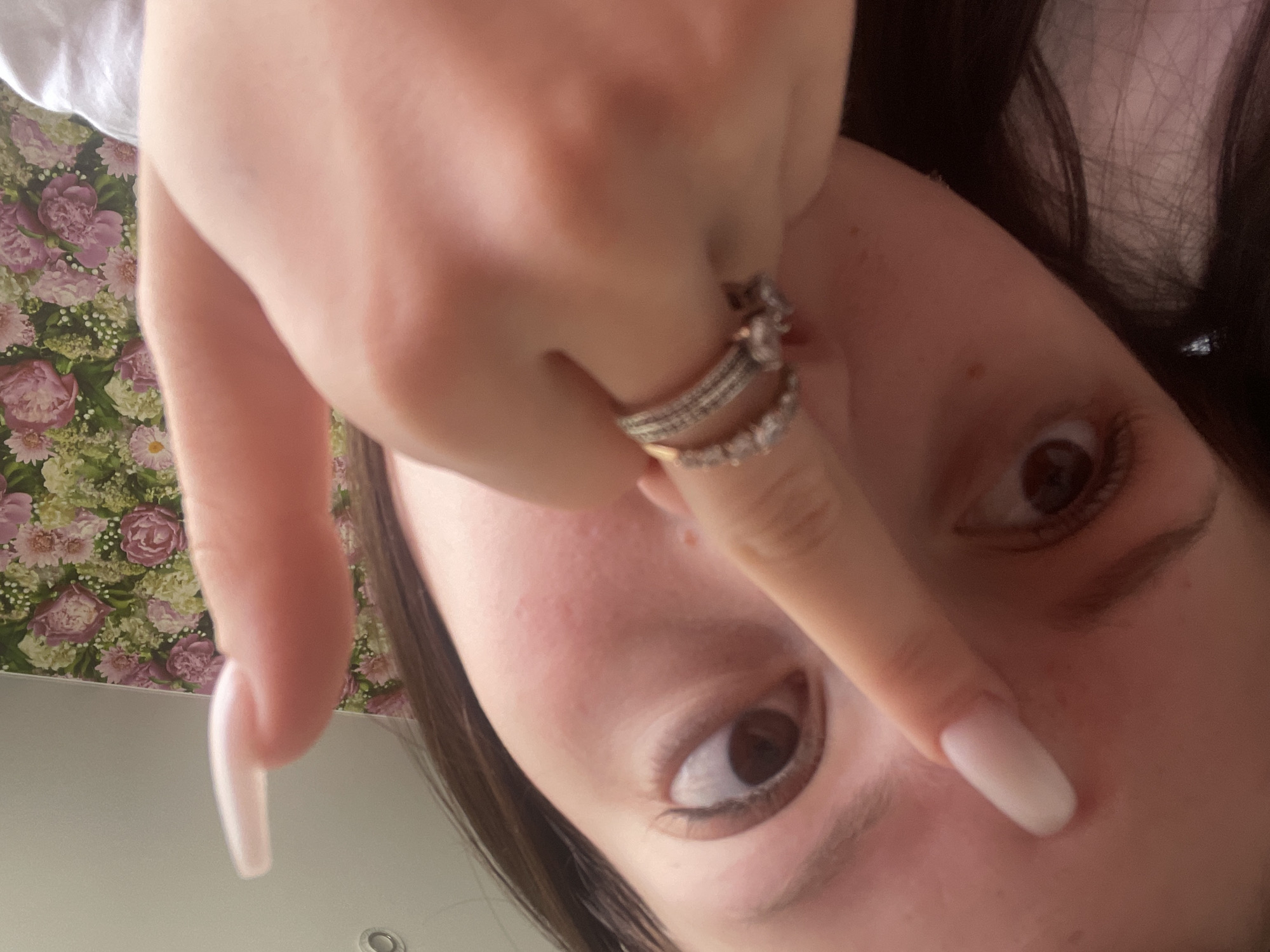 The image presents a close-up, rotated view of a person's face and hand, captured from a low angle looking upwards. The central subjects are the person's expressive brown eyes, which are wide open and directly engaging the camera, and their right hand. This hand is partially covering the upper part of the face, with the index finger touching the bridge of the nose. The fingers are adorned with several rings: one finger has a stacked set of bands, while an adjacent finger wears another ring. The nails are long, manicured, and have a light, opaque pink or nude polish. Light brown hair frames the right side of the face.
The background features a busy floral pattern with shades of pink, purple, and green, suggestive of wallpaper or upholstery. Below this, on what appears to be a ceiling, is a plain, light-colored surface with a circular recessed light fixture visible. The overall composition suggests a casual, perhaps playful, selfie or portrait taken indoors. No discernible text is present in the image. The time of day is not indicated, but the lighting appears to be ambient indoor light. The provided location context is Медведево, Russia.