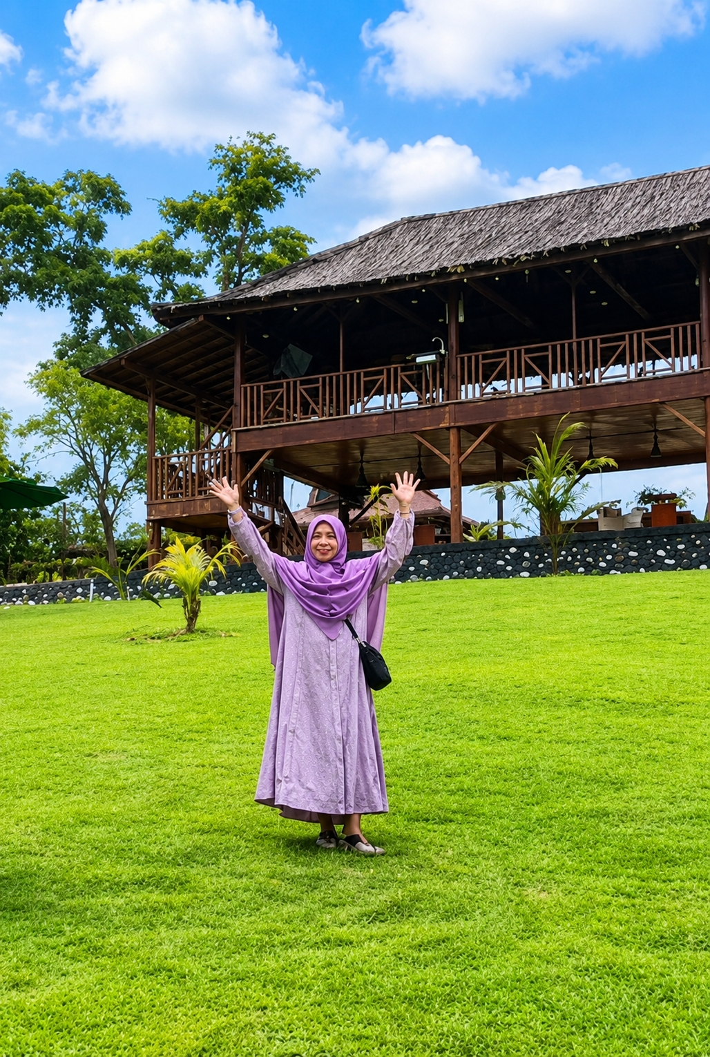 In Eampat Kwalamencirim, Indonesia, a woman wearing a purple hijab and dress stands in a grassy field with her arms raised. A wooden building with a thatched roof is visible in the background, along with trees and a blue sky with white clouds. The woman is smiling and appears to be enjoying the outdoors.