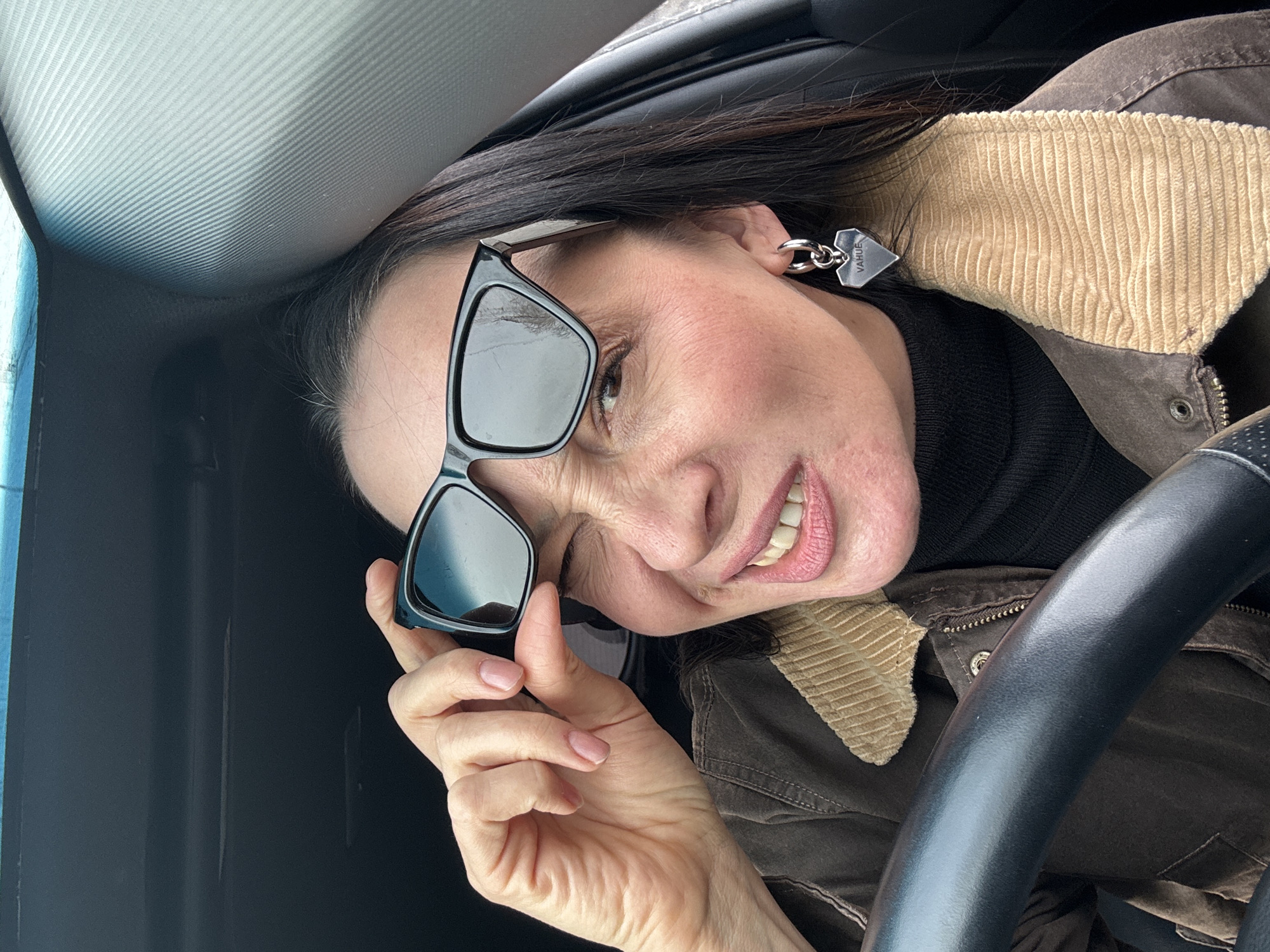 A female individual is depicted in the driver's seat of a vehicle, looking directly at the camera. She has dark hair with some visible grey strands and is wearing a dark brown jacket with a tan corduroy collar over a black turtleneck. Her left ear displays a silver-colored hoop earring with a heart-shaped charm engraved "VAHUE". She holds a pair of black-framed, dark-lensed sunglasses with her right hand, positioned over her right eye. Her left eye is closed in a wink, and her mouth is slightly open in a smile, exposing her teeth. The vehicle's interior includes a black steering wheel partially visible in the lower left, a dark headliner above, and a grey, diagonally ribbed panel on the right side, likely an A-pillar. Reflections in the sunglass lenses show an outdoor environment with sky and tree silhouettes. The precise location within the city of Chaikovsky, Russia, cannot be visually determined from the image.