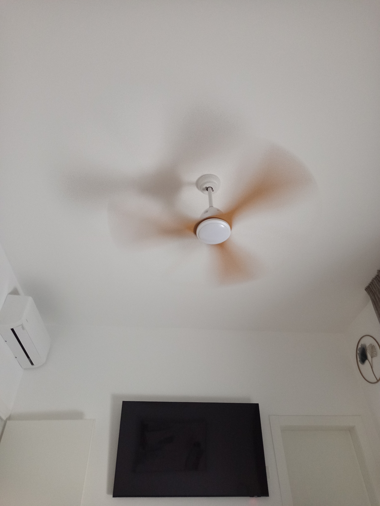 This is a low-angle, indoor photograph taken from a room in Kiryat Ata, Israel, looking upwards towards the ceiling and a portion of the walls. The primary focus is a white ceiling fan with illuminated blades, which appears to be in motion, casting a diffused shadow on the ceiling.
On the left wall, an outdoor air conditioning unit is mounted. Below it, two white, flat surfaces are visible, possibly furniture or cabinetry.
Centrally positioned on the lower wall is a large, black flat-screen television. To the right of the television, a white door frame is visible. Further to the right, near the top right corner of the image, a decorative circular wall ornament with a metallic frame and a dark floral design is present. The room appears to be brightly lit, with natural light likely coming from an unseen window or opening. The overall impression is of a clean, modern living space.