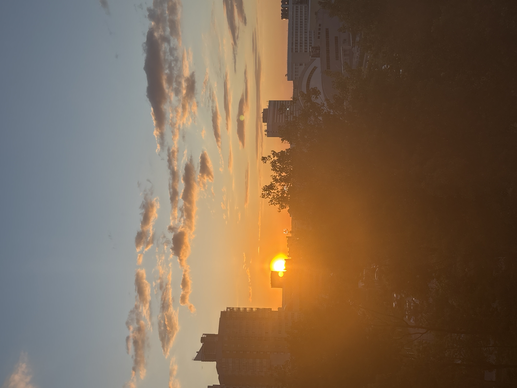 The image shows a sunset over Minsk, Belarus. The sun is low on the horizon, partially obscured by buildings and trees, creating a strong golden glow. The sky is a gradient of blue to orange, with elongated clouds illuminated by the setting sun. The foreground consists of buildings and tree silhouettes, some of which are heavily obscured by haze. The overall scene is hazy, typical of sunsets viewed over a city with potential air pollution or humidity. The image is oriented vertically.