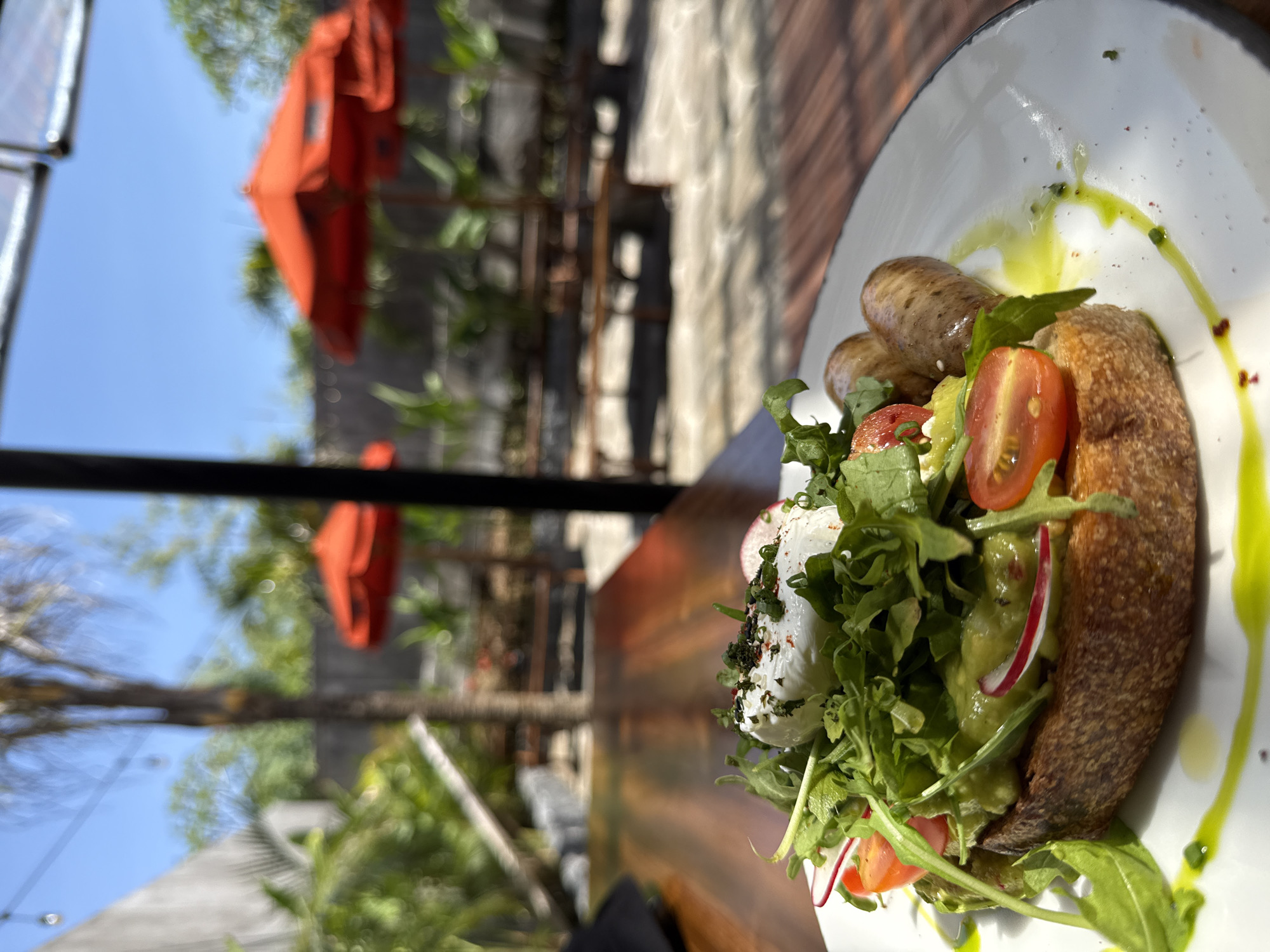 A plate of food is shown on a wooden table. The food consists of sausages, bread, a poached egg, arugula, radishes, tomatoes, and what appears to be avocado, drizzled with a green oil. The background shows an outdoor patio with orange umbrellas and greenery. The location is likely Kuta Selatan, Indonesia.