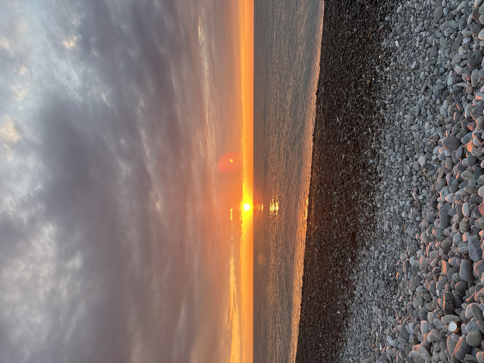 The image depicts a sunset view at a beach, likely in Batumi, Georgia. The sky is filled with clouds, tinged with shades of orange and gray from the setting sun. The sun itself is visible on the horizon, casting a bright reflection on the surface of the water. The beach is covered with pebbles, and the edge of the water is visible where the waves meet the shore. There are no visible people in the scene. The overall environment appears to be tranquil and peaceful. The photo is oriented vertically.