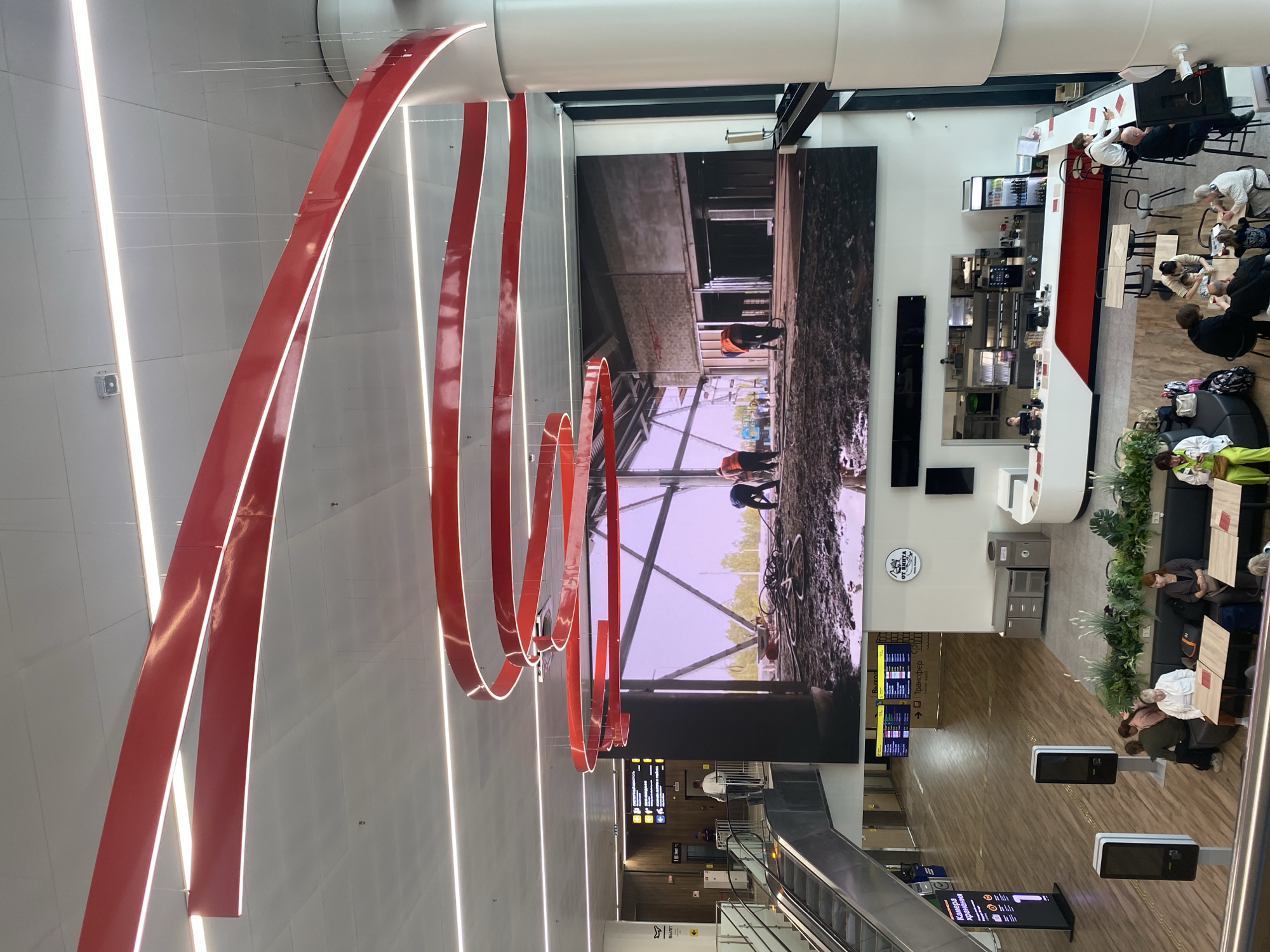 The image captures an interior scene, likely an airport terminal, with a large LED screen dominating the central wall displaying a construction site with workers. Red, curved lighting fixtures are suspended from the ceiling, adding a modern aesthetic. Below, a cafe area is visible with patrons seated at tables and a counter. Escalators and signage for departures are present, indicating a travel hub. The floor appears to be made of wood-like tiles. The overall environment suggests a public space with ongoing activity. The specific location of Novoye Martyanovo, Russia is not explicitly indicated by any visual cues within the image.