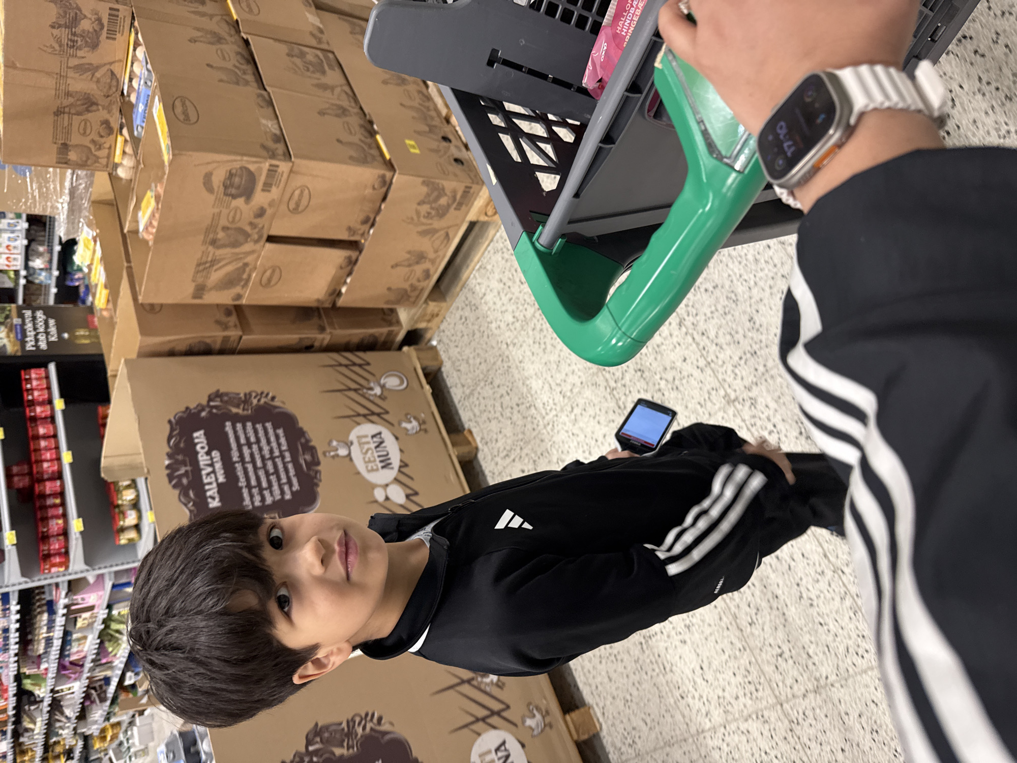 A young boy is standing in a supermarket in Tallinn, Estonia, holding a smartphone and looking at the camera. He is wearing a black Adidas tracksuit with white stripes on the sleeves and legs. Behind him, there are large cardboard boxes filled with eggs, with images of chickens printed on them. The boxes are stacked on wooden pallets. In the foreground, a shopping cart with a green handle is visible. A person's wrist wearing an Apple Watch is also visible in the foreground, resting on the shopping cart. The shelves of the supermarket are visible in the background, stocked with various products. The floor is tiled with small, speckled tiles.