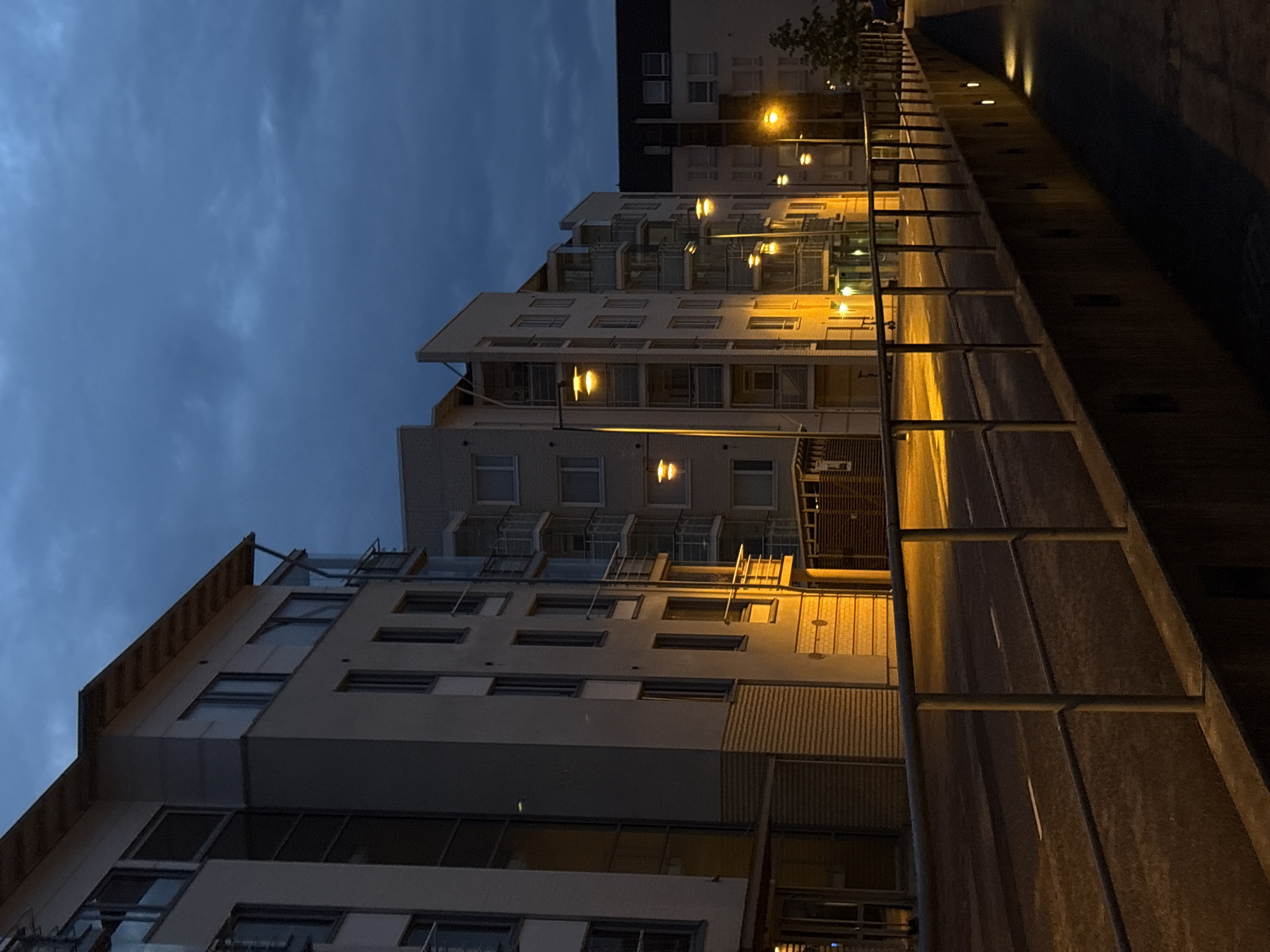 This image depicts a street scene in Pori, Finland, during twilight. The foreground features a paved walkway with a concrete barrier and a metal railing. A street runs alongside, marked with white lines and illuminated by streetlights. On the left side of the street are modern apartment buildings with multiple floors, balconies, and large windows. The buildings are lit from streetlights and interior lights, casting a warm glow. The sky above is a deep blue with visible clouds, indicating evening or impending night. The overall environment suggests a residential or mixed-use urban area. There are no people or vehicles visible in the frame.