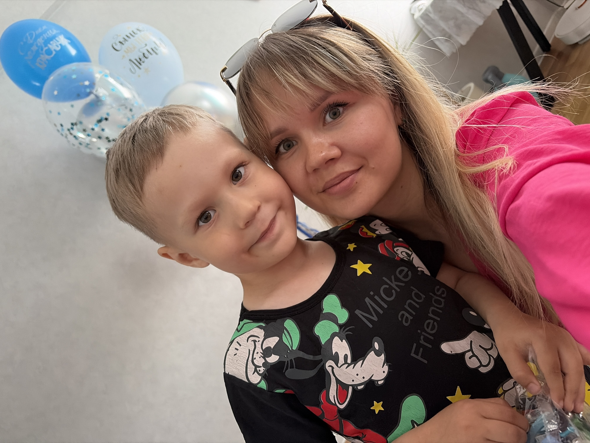 A head and shoulders selfie shows a young boy with blond hair and brown eyes posing next to a woman with blond hair and brown eyes. The boy is wearing a black t-shirt with Mickey Mouse characters and the words "Micke and Friends" printed on it. The woman is wearing a bright pink top and sunglasses on her head. In the background, there are several balloons, including blue, light blue, and clear balloons with confetti. The scene appears to be indoors, possibly a home, given the plain wall in the background. No specific location in Chaikovsky, Russia is identifiable from the image.
