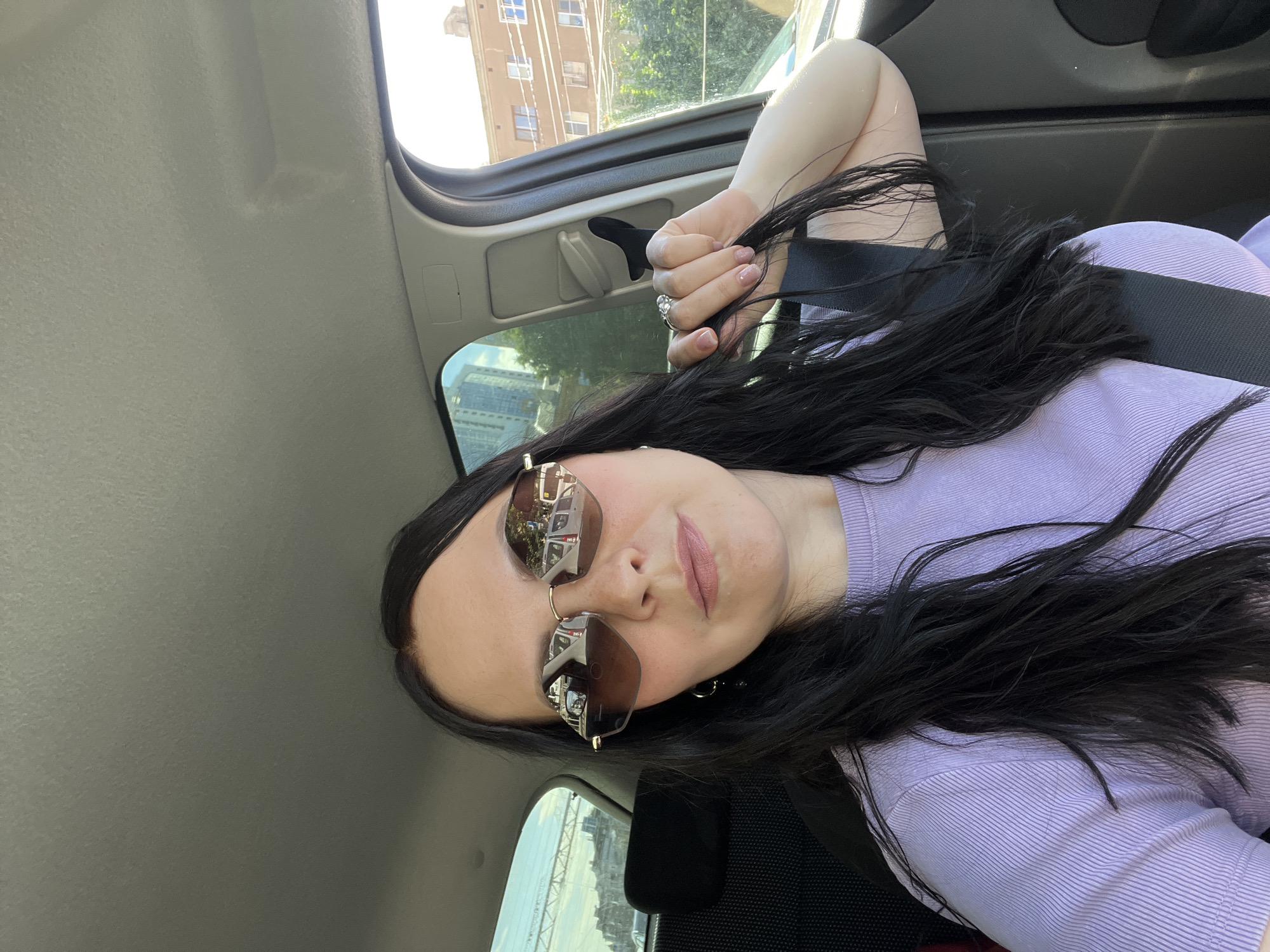 In this selfie taken from inside a car, a woman with long, dark hair is the central subject. She is wearing large, stylish sunglasses with brown lenses that reflect the scene outside, including buildings and what appears to be a white bus or van. Her lips are colored with a muted pink lipstick, and she has a ring on her right hand, which is resting on her hair. She's also wearing a lavender, ribbed top and a seatbelt. The scene suggests a bright, sunny day, possibly late morning or afternoon, given the lighting. The background visible through the car windows includes buildings and some greenery, indicating an urban or suburban setting. The overall mood is casual and relaxed, consistent with a passenger enjoying a car ride. The image provides a close-up perspective of the woman's face and upper body, with the car's interior and the outside environment offering context. There is no visible text in the image.