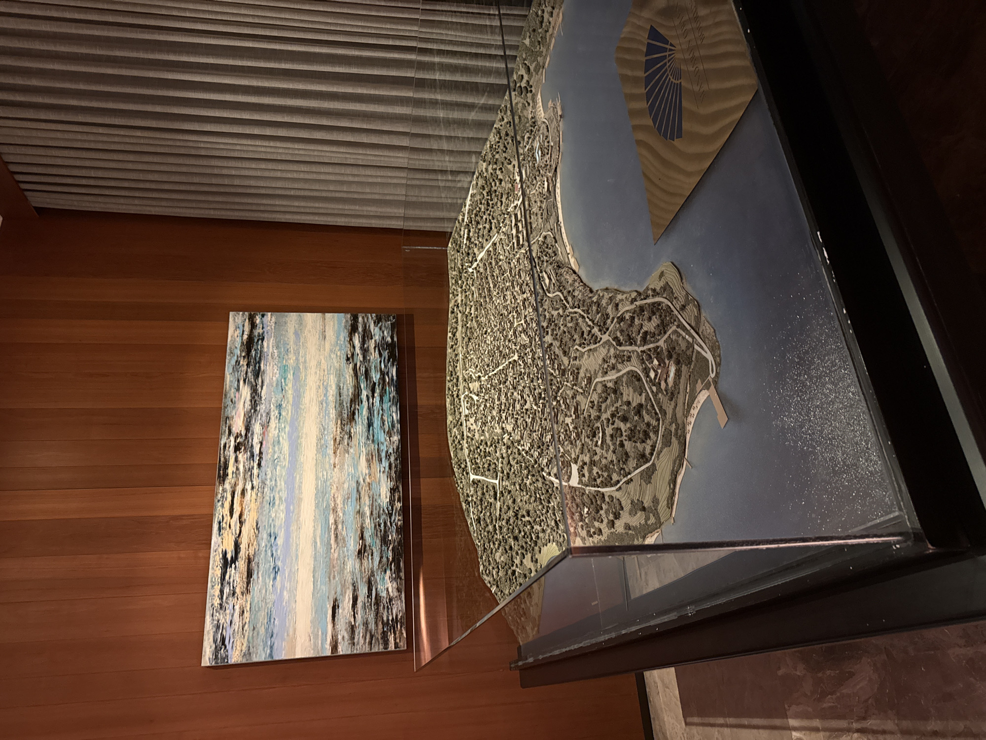 The image shows a diorama of a coastal town, likely a model of Bodrum, Turkey, displayed inside a glass case. The diorama depicts a detailed town layout with buildings, roads, and lush greenery surrounding a body of water. The town is situated on a peninsula that juts into the water, with a bay area featuring a small dock. A piece of artwork, an abstract painting with blues, whites, and black strokes, is hung on the wood-paneled wall behind the diorama. To the right of the diorama, grey, pleated curtains are visible. In the foreground of the diorama's water, a wooden coaster with a blue fan logo and the text "MANDARIN ORIENTAL BODRUM" is placed. The lighting in the scene is dim, suggesting an indoor environment, possibly a hotel lobby or exhibition space. There are no people visible in the image.