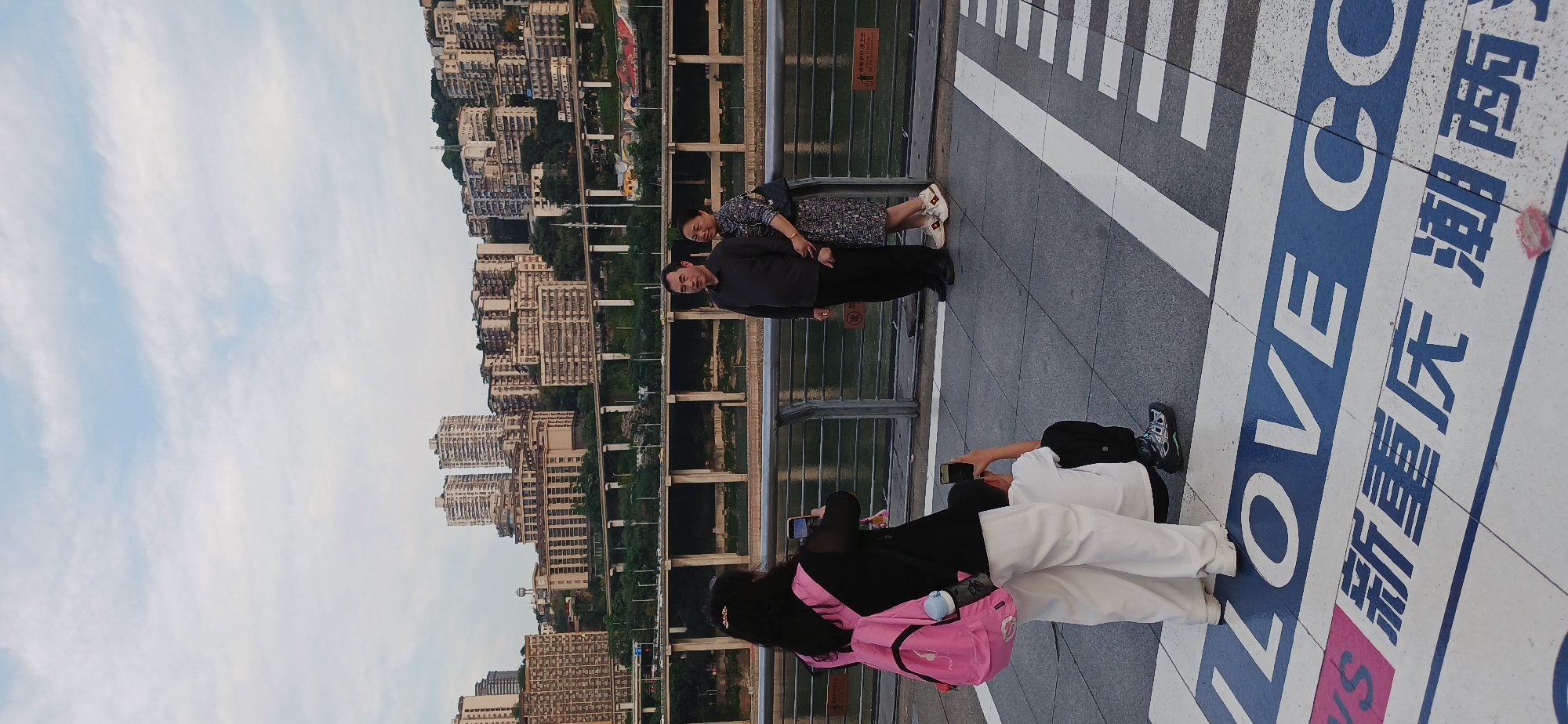 The image captures a scene in Yuzhong District, China, with a view of the cityscape and a river. A couple stands near a railing, possibly overlooking the river or a bridge. In the foreground, two individuals are seen using mobile phones. One person appears to be taking a photograph of the couple or the scenery. The pavement has a prominent design or text that includes the word "LOVE" in large blue letters along with some Chinese characters. The general environment suggests an urban setting on an elevated walkway or bridge.