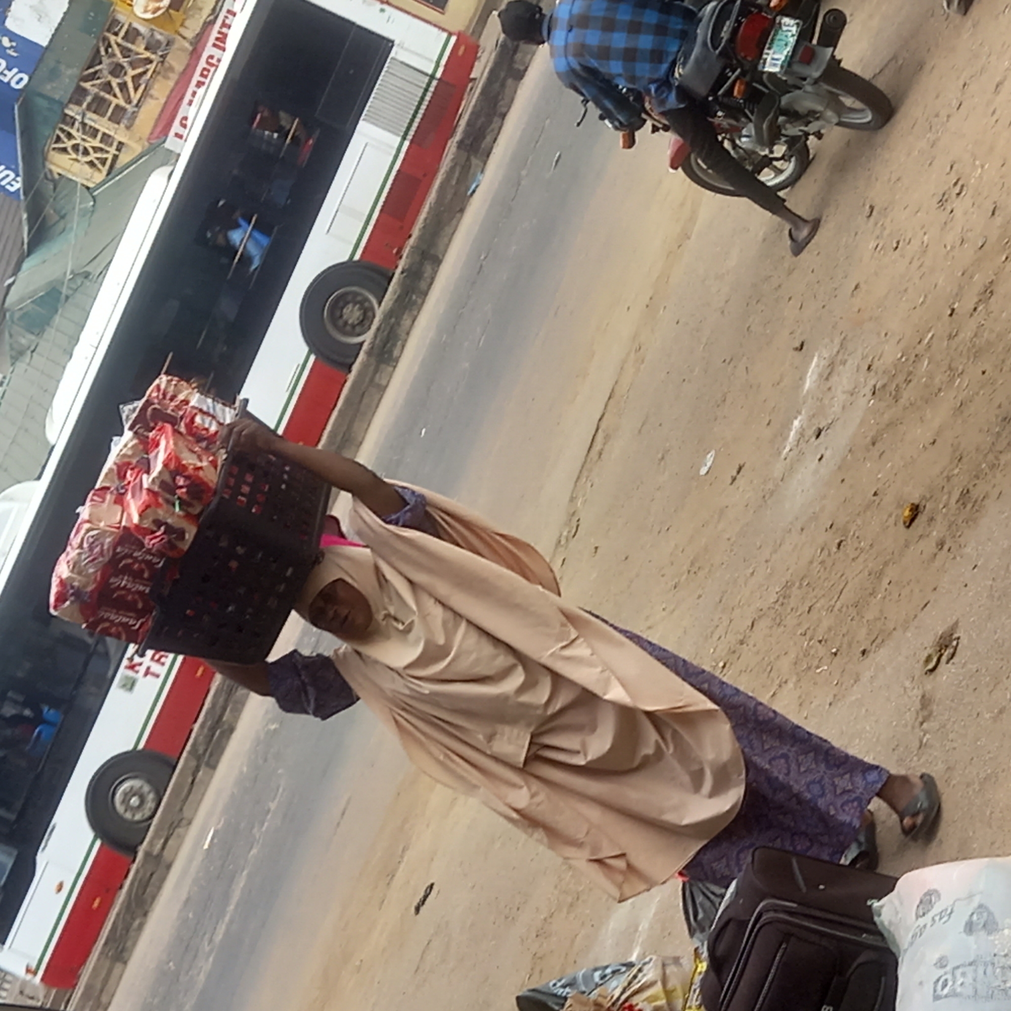 A woman in a tan hijab and blue patterned dress walks towards the right, carrying a black plastic crate filled with packages on her head. A bus is parked on the road to the left. A motorcyclist in a blue plaid shirt is on the ground on the right, partially behind the woman. Suitcases and bags are on the ground in the lower right. The location is Sardauna, Nigeria.