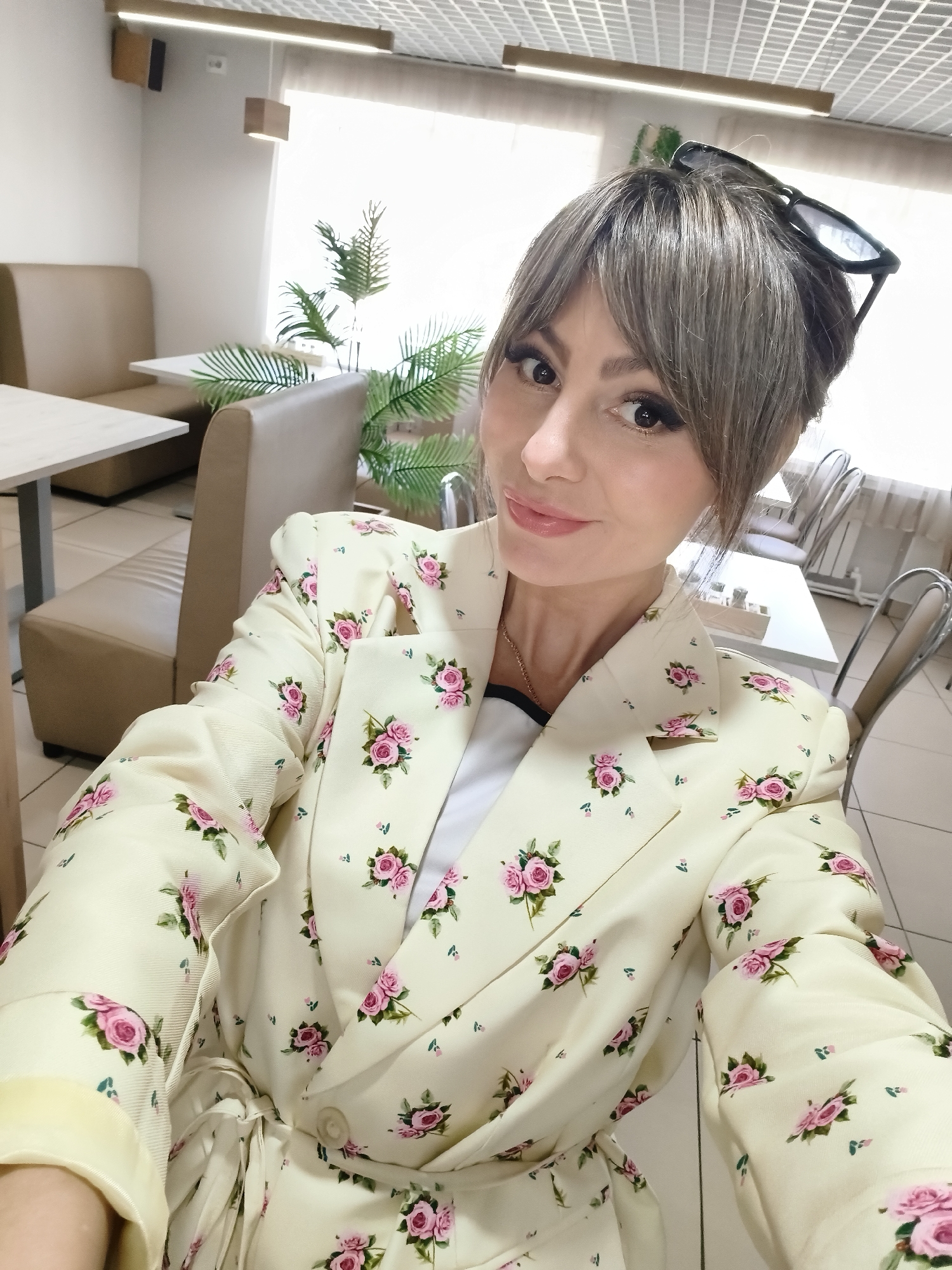 The image shows a woman in a floral blazer taking a selfie. She has light skin, brown eyes, and bangs. Sunglasses are perched on top of her head. She is wearing a yellow blazer with small pink rose patterns, possibly with a tie belt at the waist. Underneath the blazer, a white top with a black trim is visible. The background suggests she is in a cafe or restaurant. There is booth-style seating with beige upholstery and tables with white tops. A potted plant is near the window, which provides natural light. Metal chairs with white seats are also visible in the background. The location is Chaikovsky, Russia.