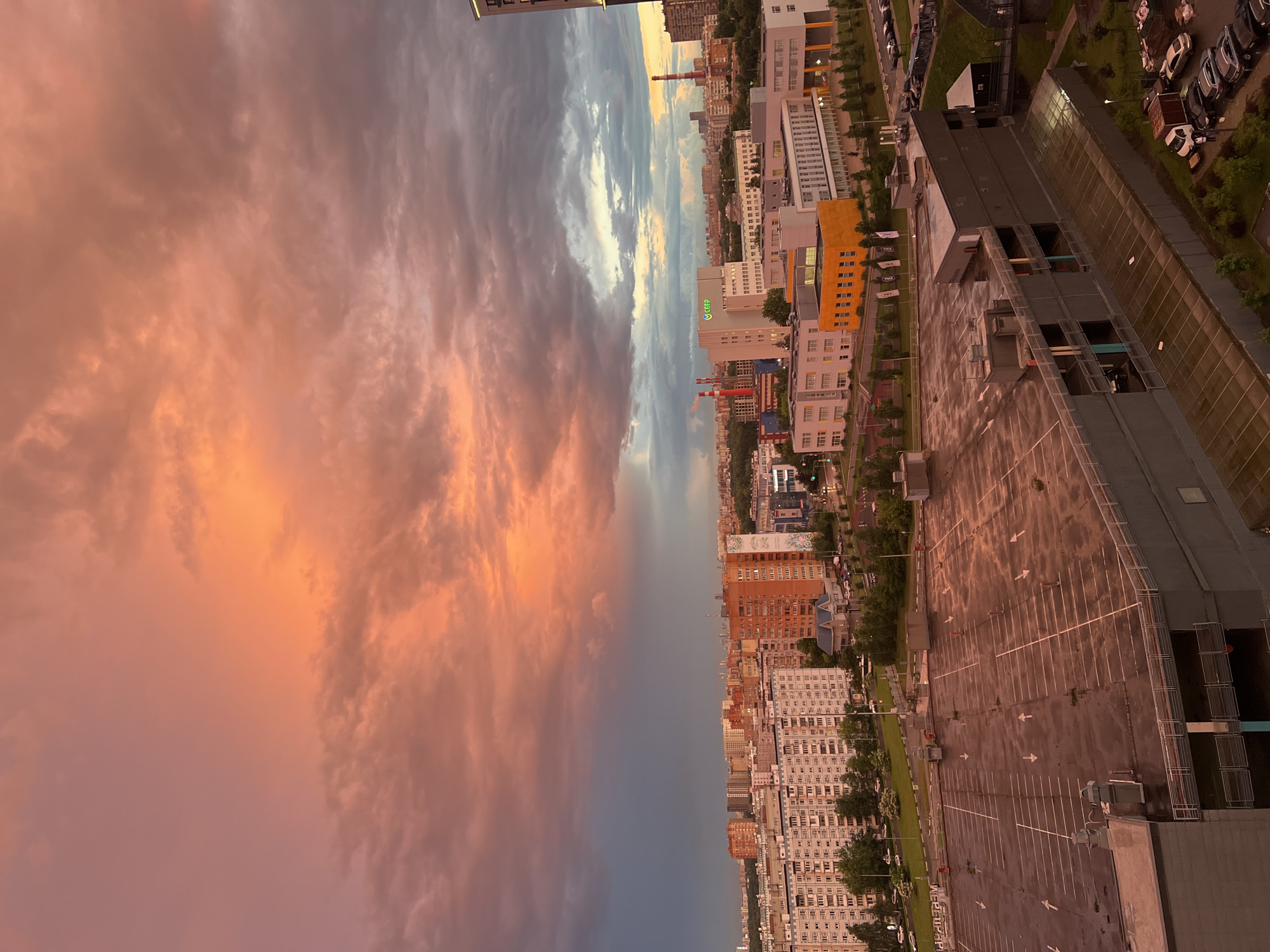 This is a landscape orientation, low-angle aerial shot of a city skyline at sunset. The sky is filled with dramatic clouds, colored in shades of orange, pink, and grey, suggesting a vibrant sunset. To the right of the frame, a sprawling cityscape is visible, characterized by numerous tall residential buildings and some commercial structures. A large, empty parking lot dominates the foreground on the right side of the image. Green trees and streetlights line the streets between the buildings and the parking lot. The overall scene depicts an urban environment at dusk, with a focus on the interplay between the natural beauty of the sunset sky and the man-made structures of the city. The image evokes a sense of twilight and urban tranquility. There is no visible text in the image.