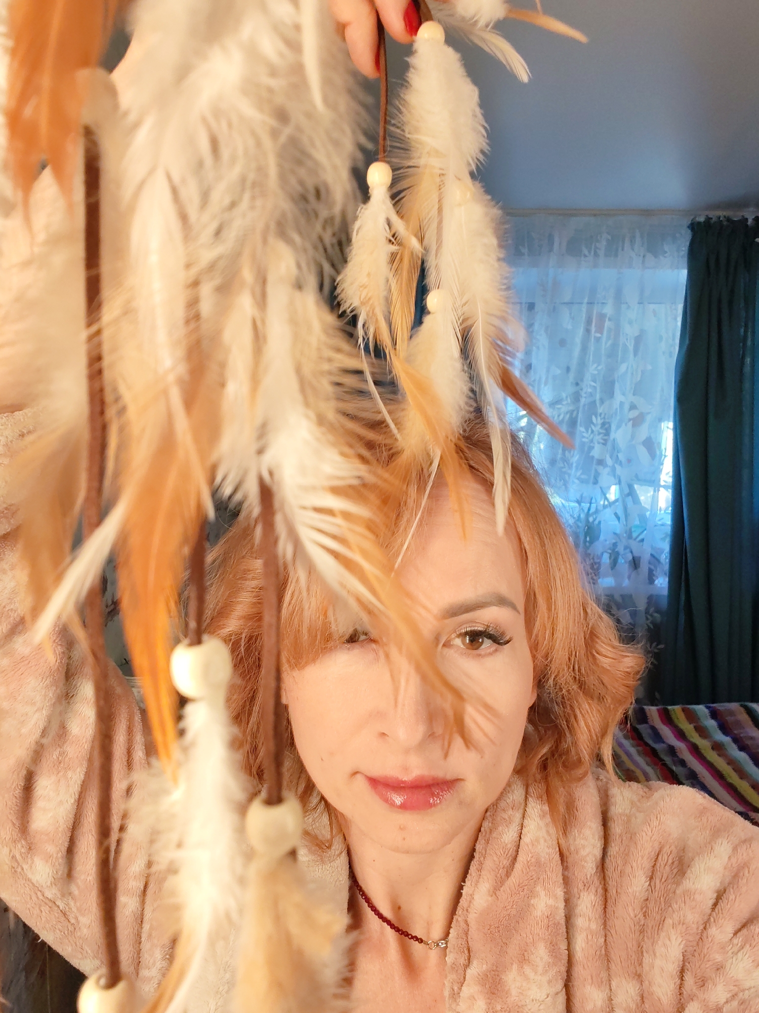 A fair-skinned woman with ginger hair, wearing a pink fuzzy bathrobe and a maroon bead necklace, is holding a dreamcatcher with white and brown feathers. She is looking directly at the camera with a slight smile. The background shows a window with sheer white curtains and teal drapes, and a glimpse of a multicolored striped surface. The location is assumed to be Chaikovsky, Russia.