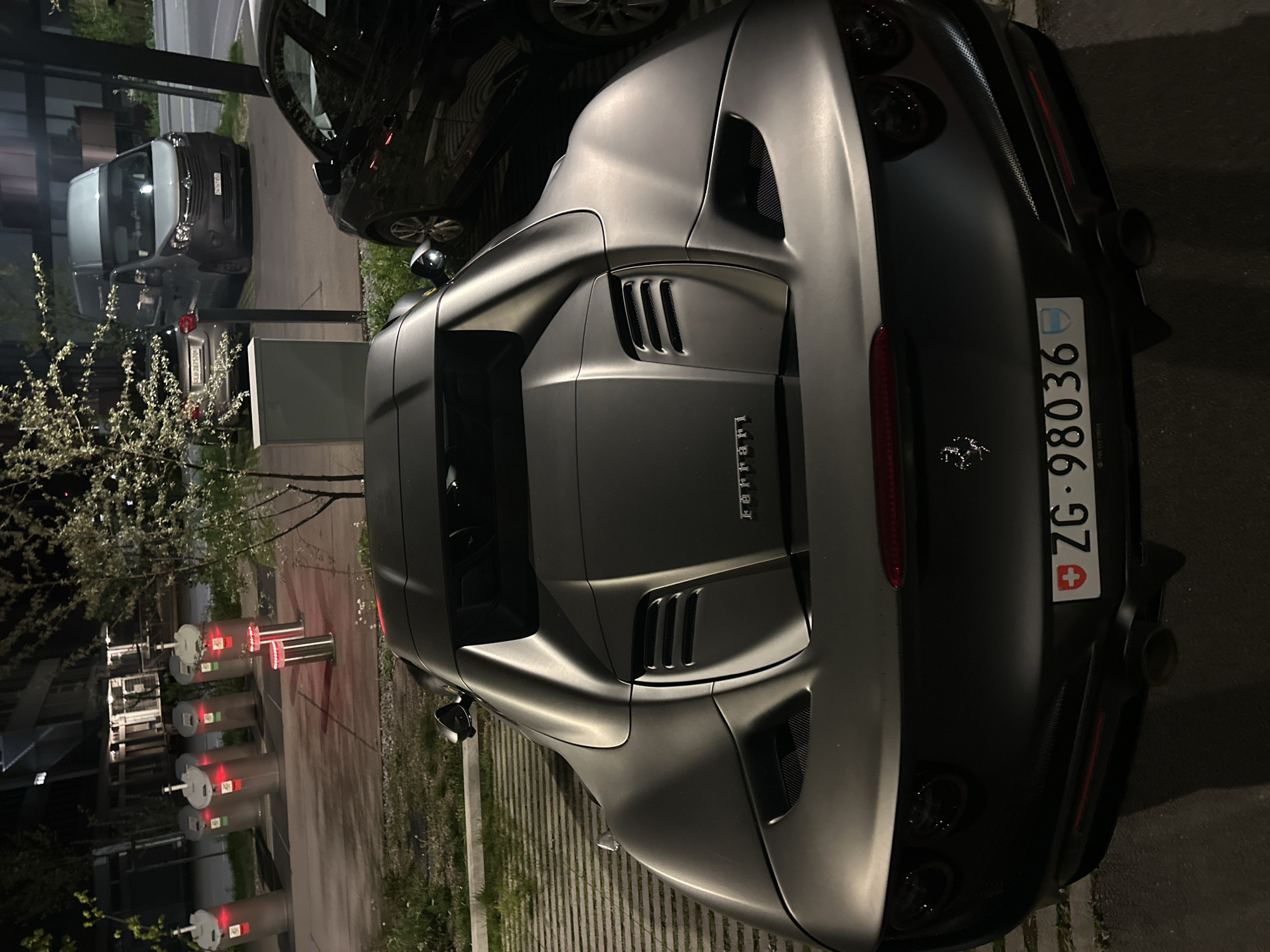 The image displays a matte dark gray Ferrari sports car parked outdoors at night, viewed from the rear-right. The car's sleek lines, pronounced rear fenders, and multiple air vents on the engine cover are prominent. The word "Ferrari" is visible in chrome lettering on the trunk lid, accompanied by the iconic prancing horse emblem. The rear of the vehicle features quad exhaust tips and a red horizontal taillight bar.
A Swiss license plate, "ZG - 98036," is clearly displayed on the back of the Ferrari, indicating registration in the Canton of Zug. The Swiss flag and the Zug canton emblem are also visible on the plate.
To the left of the Ferrari, a series of modern, cylindrical bollards with illuminated red tops are arranged, suggesting an access control point. Beyond these bollards, trees or bushes with light-colored blossoms add a touch of nature to the scene. In the background, other vehicles are parked, including a dark SUV or sedan partially obscured by the Ferrari, a light-colored commercial van, and another dark sedan. The setting is a paved outdoor area, possibly a private driveway or parking lot, dimly lit by artificial lights, indicating nighttime or late evening. The location is contextualized as Dübendorf, Switzerland.