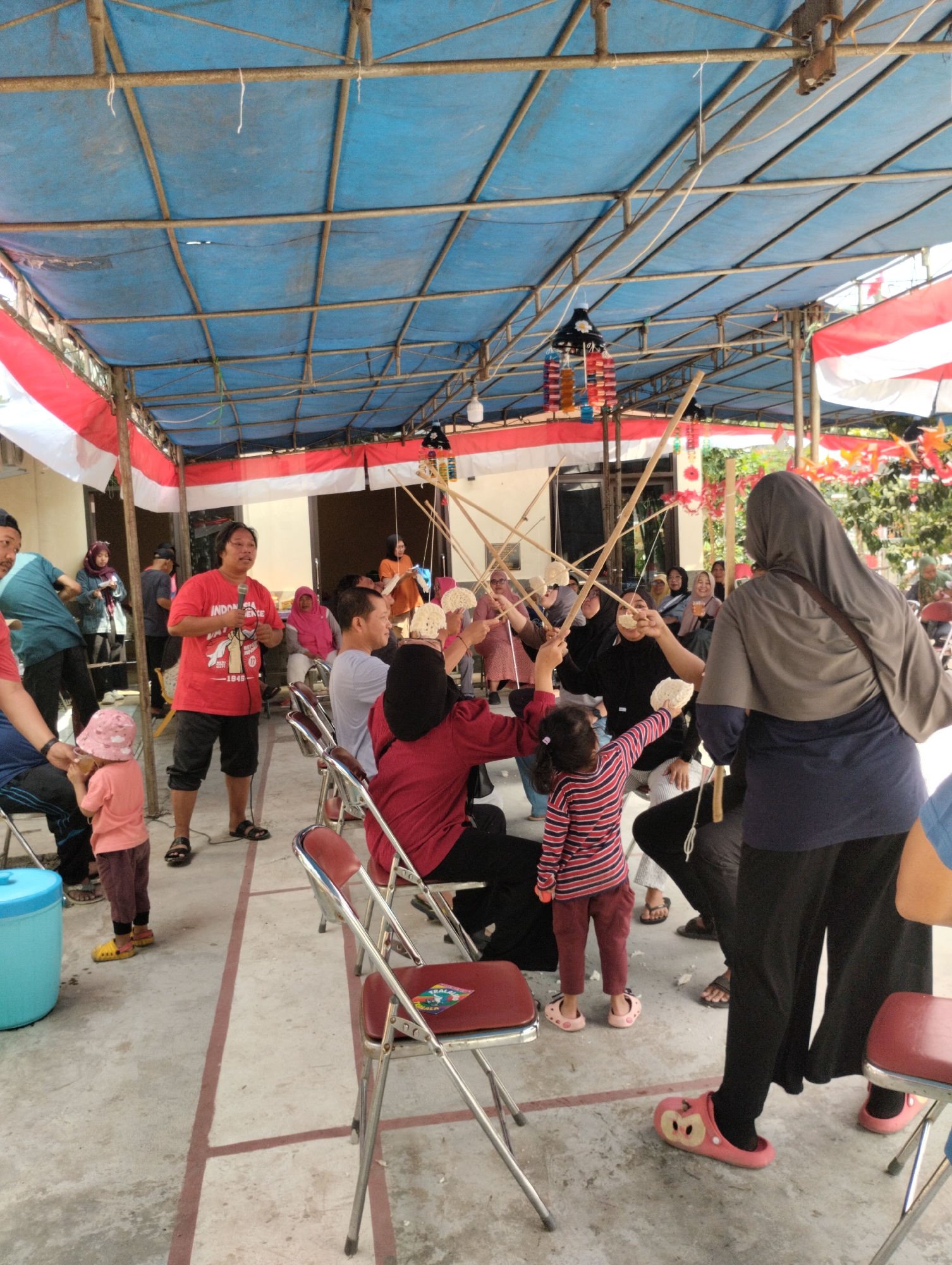 This image depicts an outdoor event, likely a community gathering or celebration, taking place in Bekasi, Indonesia. A temporary shelter, constructed with a blue tarpaulin roof supported by metal poles, covers the area. Red and white Indonesian flags are prominently displayed, adorning the sides of the shelter, suggesting a patriotic theme.
A group of people, including adults and children, are engaged in an activity involving long bamboo poles with attached food items, possibly crackers or snacks. The participants are holding these poles and attempting to grab the food, indicating a game or competition. Several individuals are seated on folding chairs, while others are standing.
In the center of the frame, a man in a red t-shirt with Indonesian text and graphics is holding a microphone, appearing to be addressing the crowd or participating in the event. Children are actively involved, with one young girl in a striped shirt reaching for food from a pole. Another child, wearing a pink hat and dress, is standing near a large blue plastic container.
The ground is paved concrete, with red lines marking sections or pathways. The background reveals buildings and vegetation, hinting at an urban or semi-urban setting. The overall atmosphere appears lively and festive.