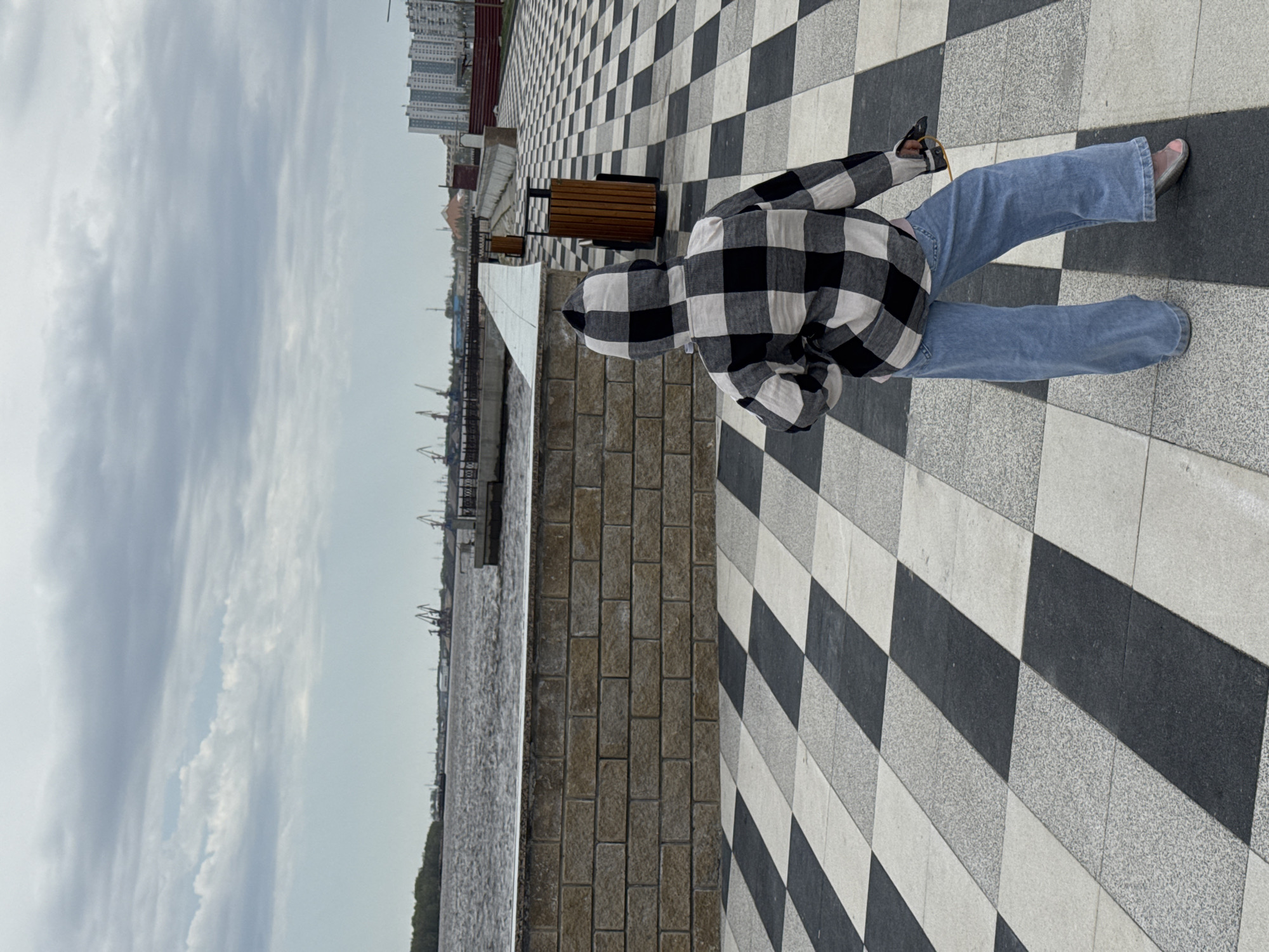 A person in a hooded, black and white plaid shirt and light blue jeans stands on a black and white checkered pavement, looking out at a river on an overcast day. To the left of the pavement is a brick wall and a body of water with cranes and other industrial structures visible in the distance. Buildings are also visible further in the background on the right. Benches are seen along the walkway. The location is Nizhnevartovsk, Russia.