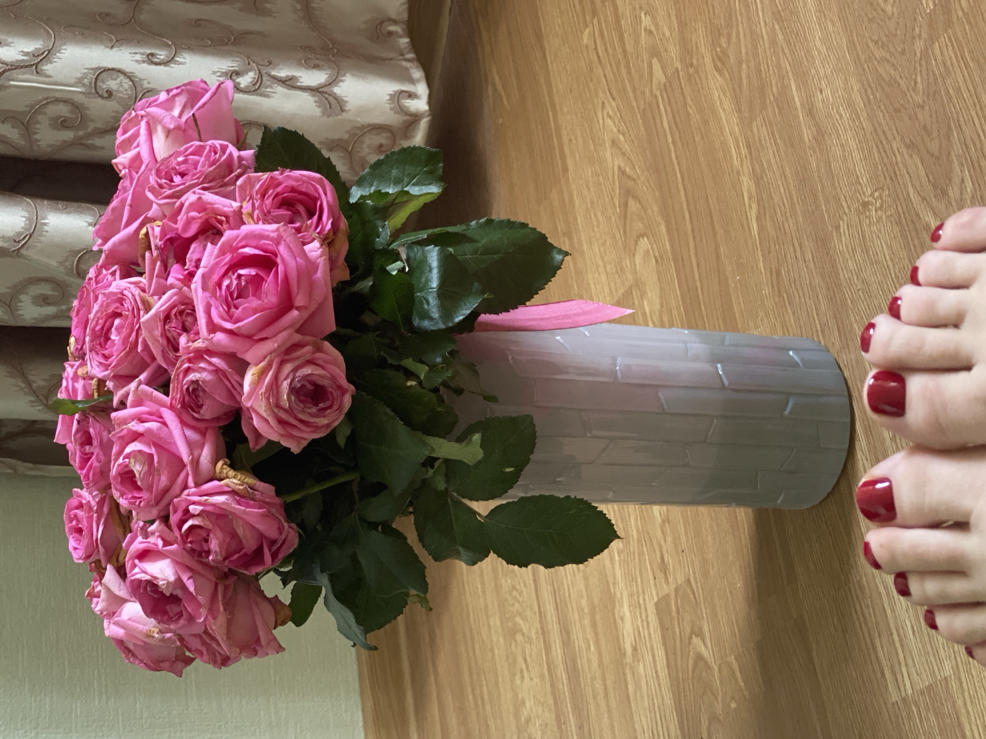 A bouquet of pink roses sits in a gray vase on a wooden floor, near a pair of feet with red nail polish. The scene is near a curtain with a swirling pattern. The location is assumed to be Sochi, Russia.