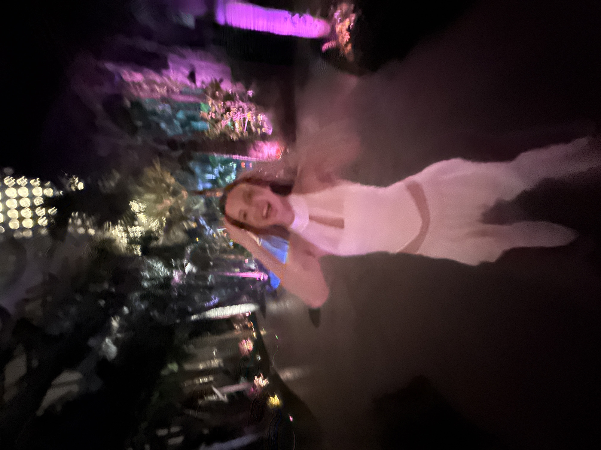 The image captures a vibrant, joyful night scene, likely in an illuminated garden or event space in Pudong, China. A young woman is the central subject, positioned towards the right of the frame, smiling broadly and looking directly at the camera. She has long, light brown hair and is wearing a light-colored, possibly white or light pink, halter-neck dress or top with a flowing skirt. Her left hand is casually placed near her head.
The background is a dynamic blur of artificial lights and dark foliage, creating a festive atmosphere. Dominant colors in the lighting include vivid purples, pinks, and greens, illuminating trees, plants, and various structures. Some lights appear as distinct, bright bokeh circles, indicating a shallow depth of field or motion blur. The setting suggests an outdoor or semi-outdoor evening event, such as a light festival, a decorated park, or a night-time garden exhibit. The time of day is clearly night, evidenced by the darkness and the reliance on artificial illumination. The woman's wide smile conveys happiness and enjoyment, suggesting she is actively participating in or attending a pleasant social event. No visible text is present in the image.
