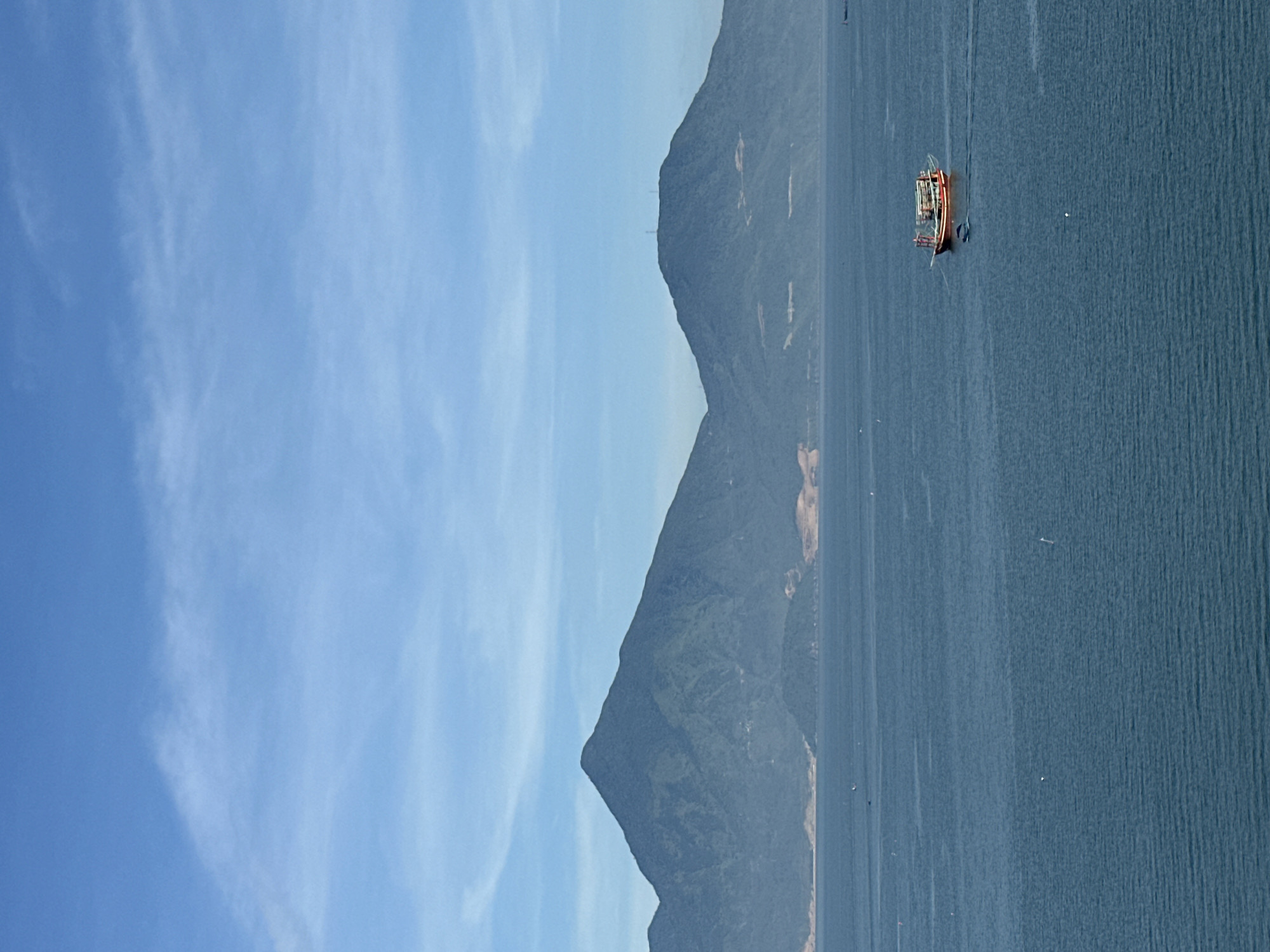 The image shows a scenic view of the sea and mountains near Đà Nẵng, Vietnam. In the foreground, the calm, blue sea fills most of the frame, with gentle ripples visible on the surface. A small, red and brown boat with a canopy is navigating the water in the middle right of the image, leaving a faint wake. Further in the distance, two large, green, mountainous islands dominate the horizon. The left mountain is tall and pointed, while the right one is more rounded with a few structures visible on its peak. The sky above is a clear, bright blue with thin, wispy white clouds scattered across it, suggesting a pleasant, sunny day. The overall impression is one of tranquility and natural beauty.