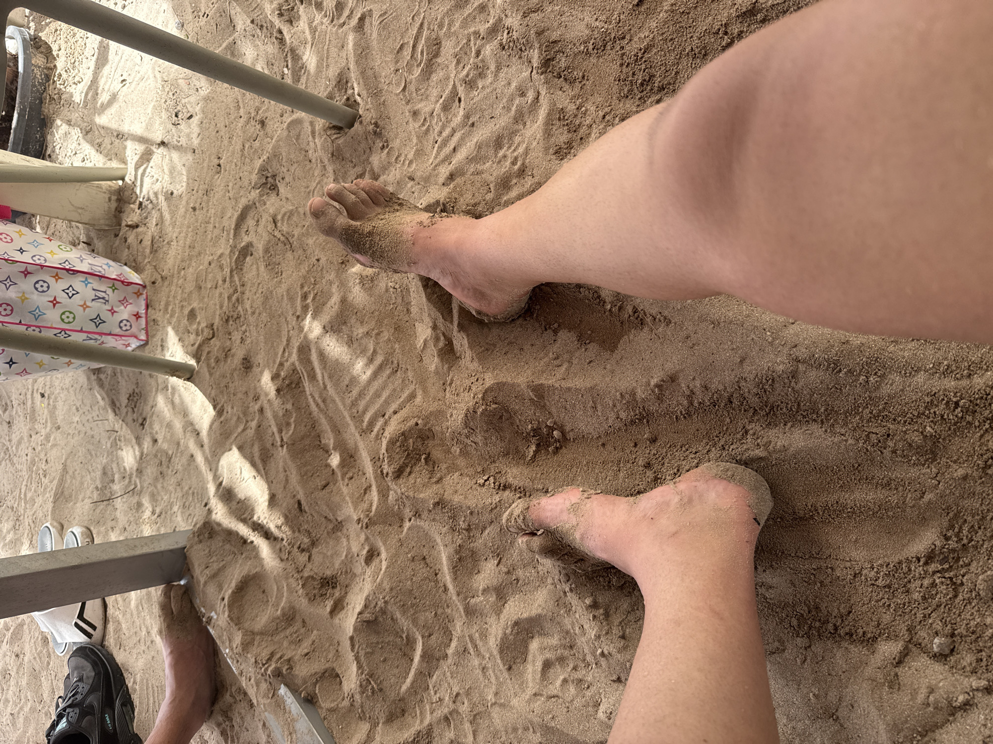 The image shows a first-person perspective of someone sitting in the sand, with their legs and feet partially visible. The sand is light brown and appears to be on a beach, likely in Platja De Muro, Spain, given the context. In the background, there are chair legs and a bag with a colorful pattern. The person's legs and feet are covered in sand. To the left, a black athletic shoe is also visible. The lighting suggests it is daytime, with shadows cast on the sand.