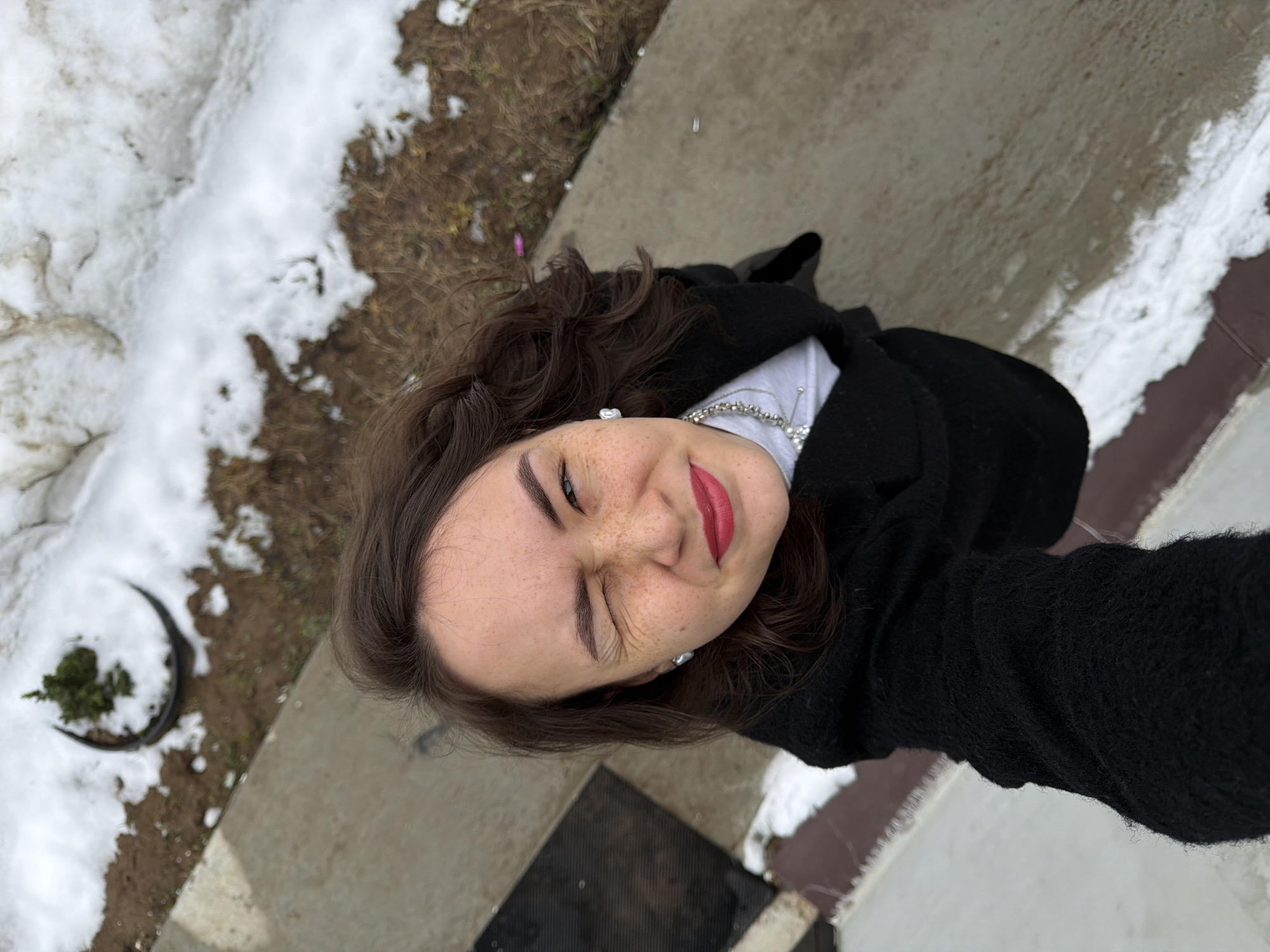 A young woman is pictured in a selfie taken outdoors. The camera is held at an angle looking up at her. She is wearing a black coat and a light-colored top with a silver necklace. The woman's right eye is squinted shut, while her left eye is open. She has brown hair with some curls and visible freckles on her face. Her lips are painted a red color.
The background shows a concrete or stone surface covered in patches of snow. There is also some dirt and sparse vegetation visible. It may be Chaikovsky, Russia.
