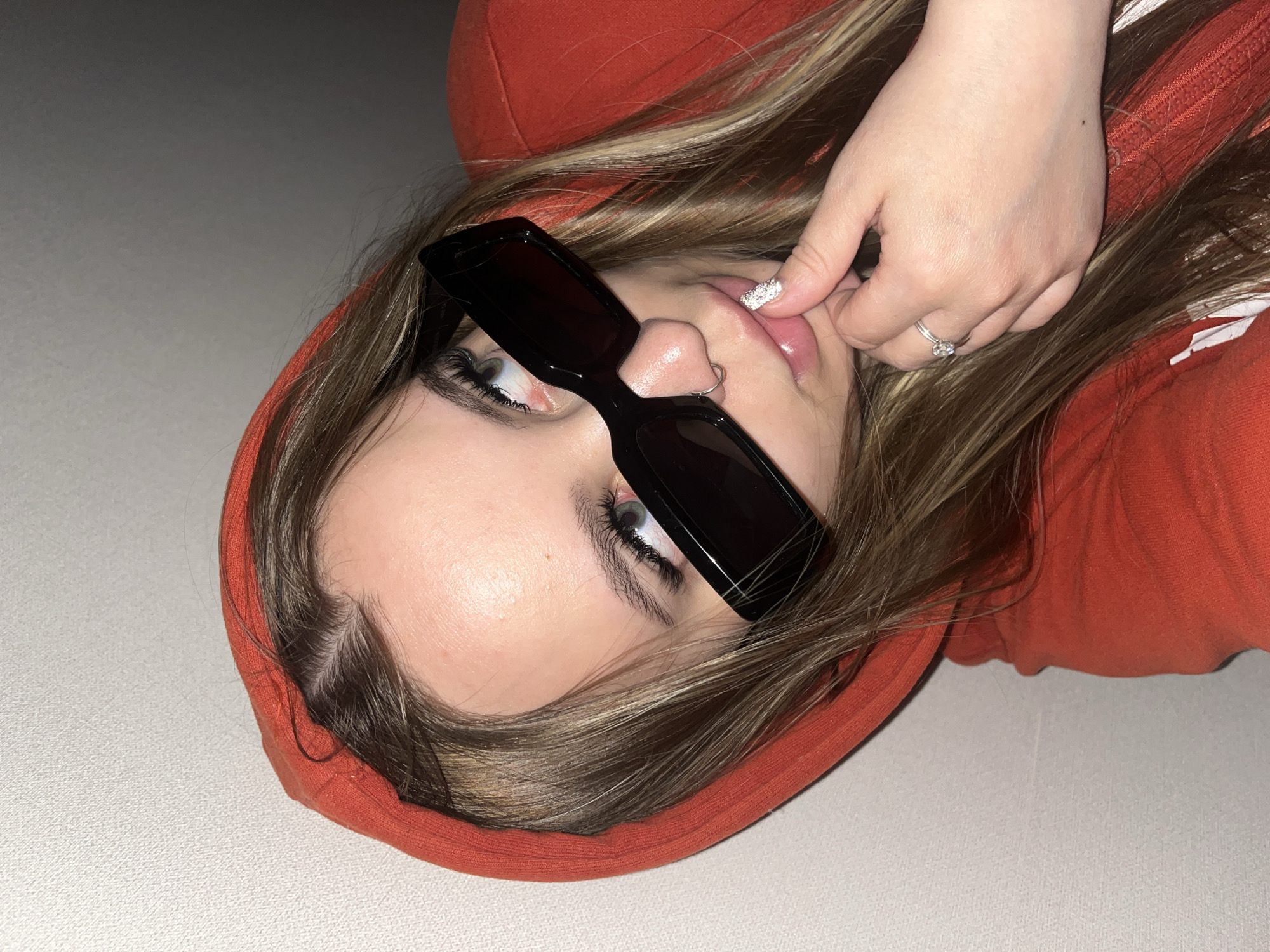 A close-up, angled shot features a young person lying down, possibly indoors, against a plain, light-colored surface. The individual is wearing a vibrant orange hooded top, with their light brown hair, streaked with lighter tones, framing their face. Black, rectangular-framed sunglasses are positioned on their nose, partially obscuring the eyes, though a light-colored iris is visible on the left eye. A small silver hoop nose ring adorns the right nostril. The person's right hand is brought up towards their face, with the index finger gently touching the lower lip. The fingernail of this finger is notable for its sparkling, silver-colored manicure. A silver ring with a clear central stone is worn on the ring finger of the same hand. The pose is casual and somewhat playful, with the subject looking towards the viewer from this angled perspective. No visible text or specific location cues are present within the image.