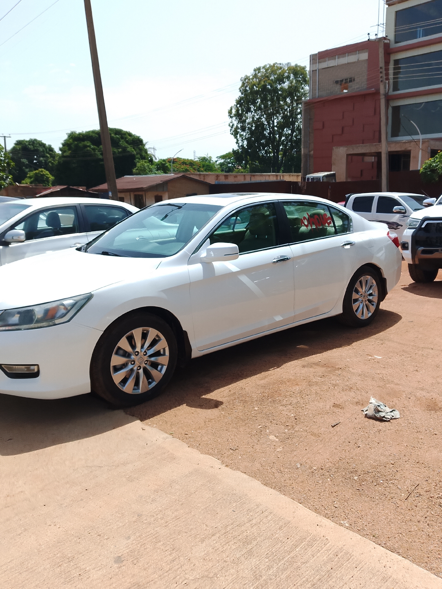 A white Honda Accord sedan is parked on a dirt lot in front of a red brick building with a modern glass facade. The car is facing left, and its front passenger side is visible. The building appears to be a commercial establishment, possibly an office or a store. Several other vehicles, including a white SUV and a black truck, are parked in the background. The sky is clear and blue. A tall wooden pole stands to the left of the car, and some trees are visible in the distance. The scene appears to be in an urban or semi-urban area in Jos, Nigeria.
