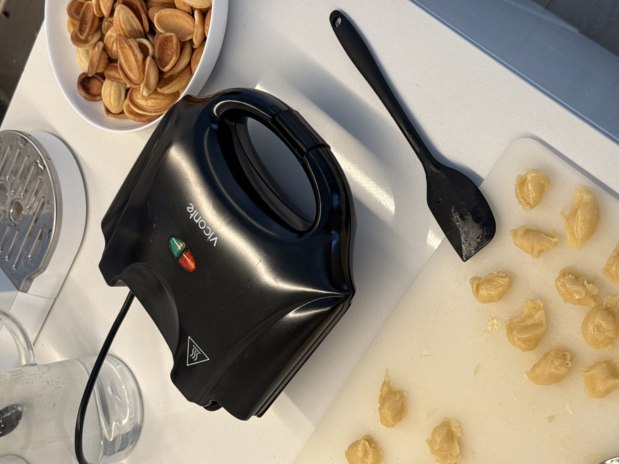 The image displays a kitchen countertop scene in Башкултаево, Russia, focused on the preparation of Oreshki cookies. A black Viconte electric appliance, designed for baking, is prominently featured, displaying both red and green illuminated indicator lights and a warning symbol for heat. To the left of the appliance, a clear glass pitcher filled with water is visible. To the right, a white bowl holds a quantity of baked, golden-brown, shell-shaped cookies. In the foreground, a white cutting board is populated with multiple small, unbaked portions of pale yellow dough. A black spatula with a perforated handle lies on the countertop near the cutting board. A segment of a larger white kitchen appliance with a metallic grater is partially visible in the upper background. No individuals are present in the frame.